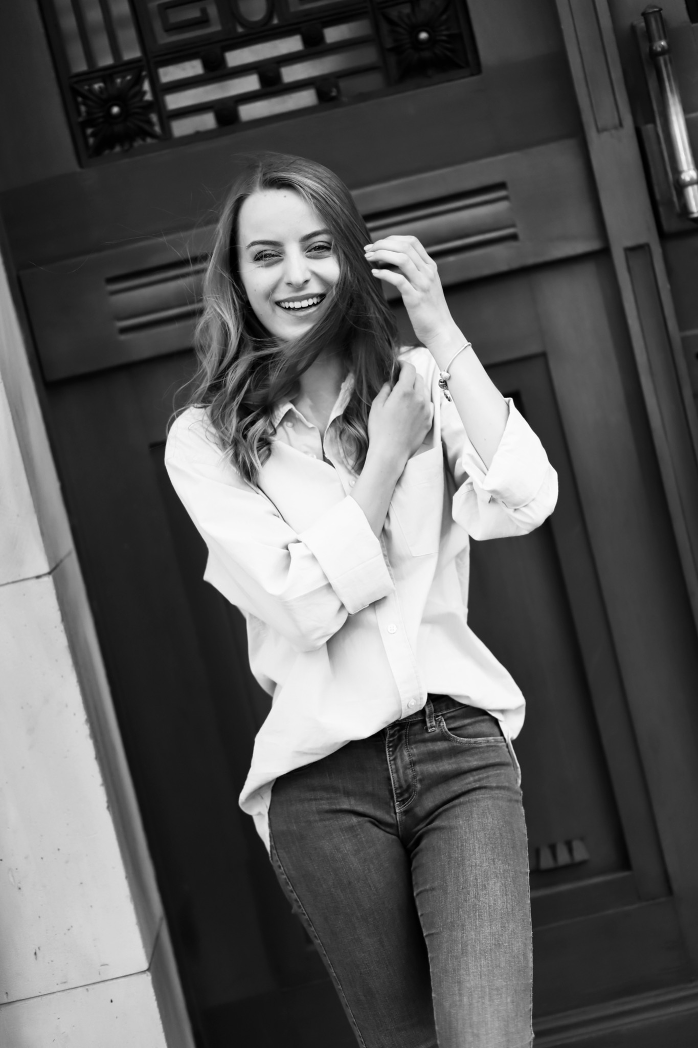 Portret uśmiechniętej kobiety w jeansach i białej koszuli, pozującej na tle ciemnych, zdobionych drzwi w czerni i bieli.