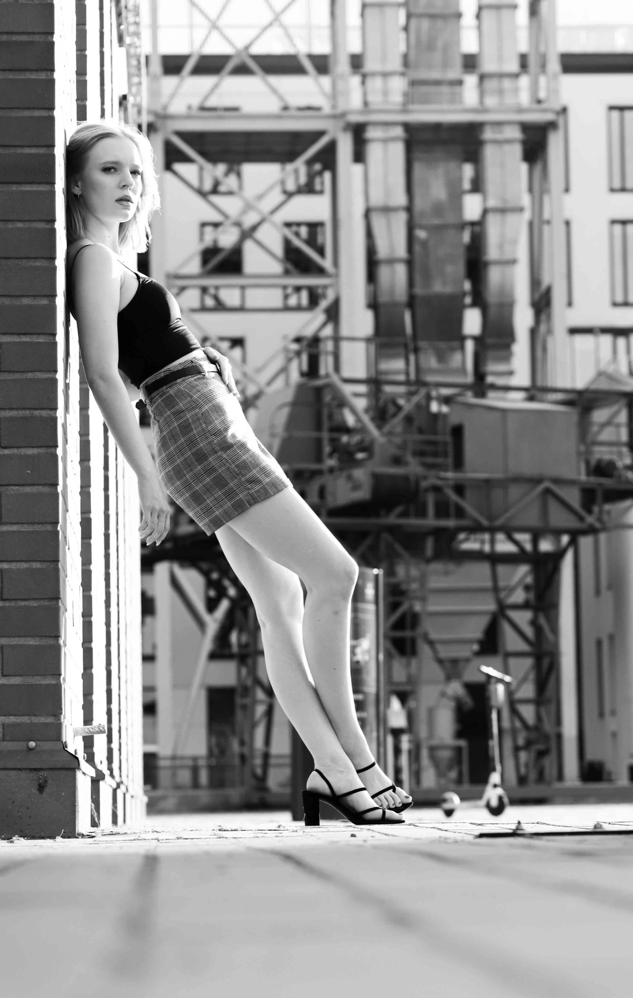 Czarno-białe zdjęcie młodej kobiety w spódnicy w kratę i czarnym topie, opierającej się o ceglaną ścianę na tle industrialnego krajobrazu z dźwigami i konstrukcjami stalowymi.
