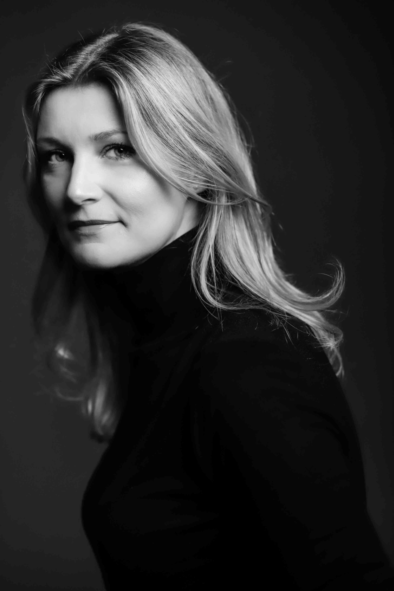 Portret kobiety o jasnych włosach, ubrana w czarny golf, na ciemnym tle, w czerni i bieli, z delikatnym uśmiechem.