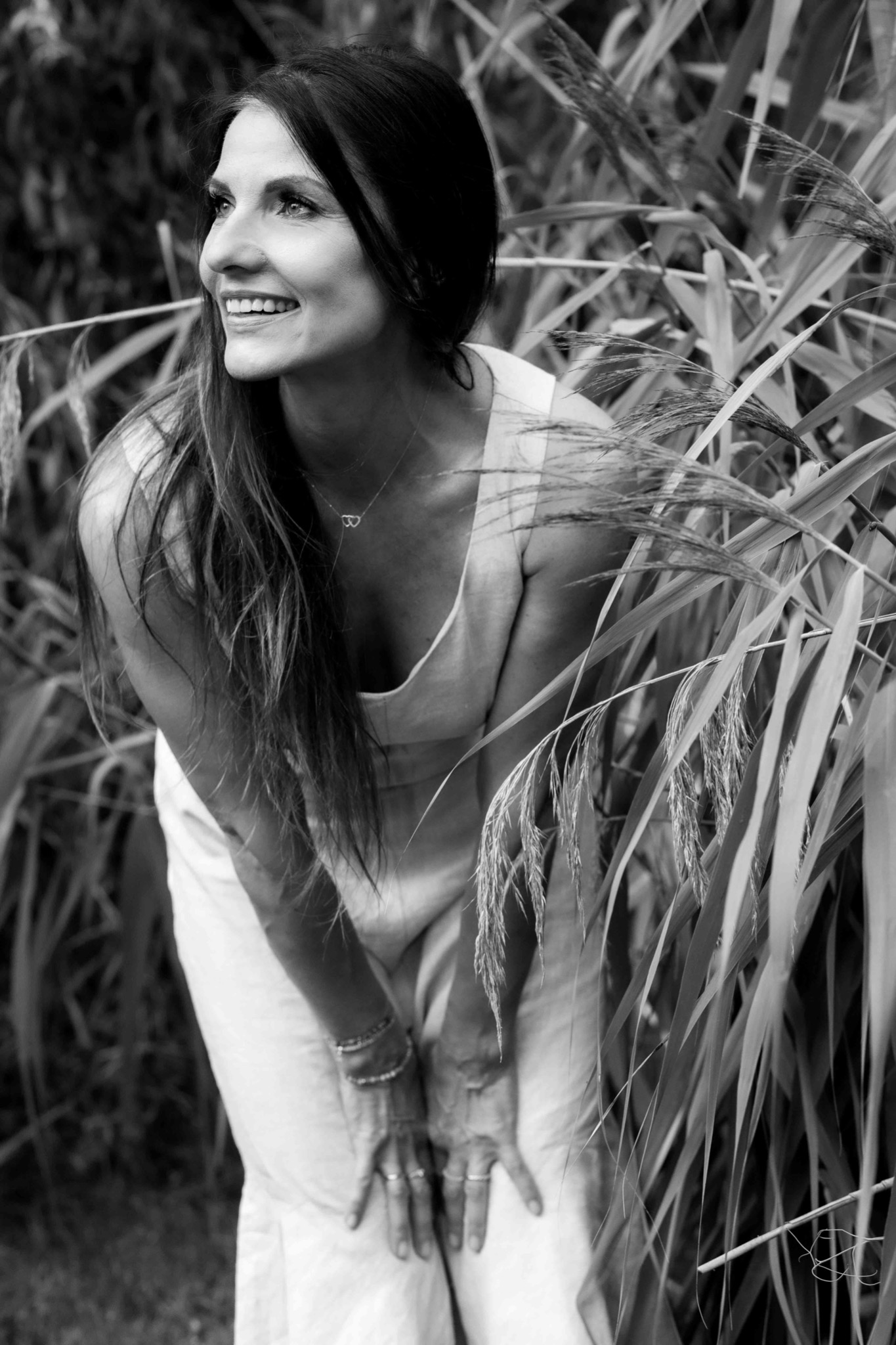Portret uśmiechniętej kobiety w plenerze, czarno-białe zdjęcie, z delikatną biżuterią i dłońmi opartymi na kolanach, otoczonej wysokimi trawami.