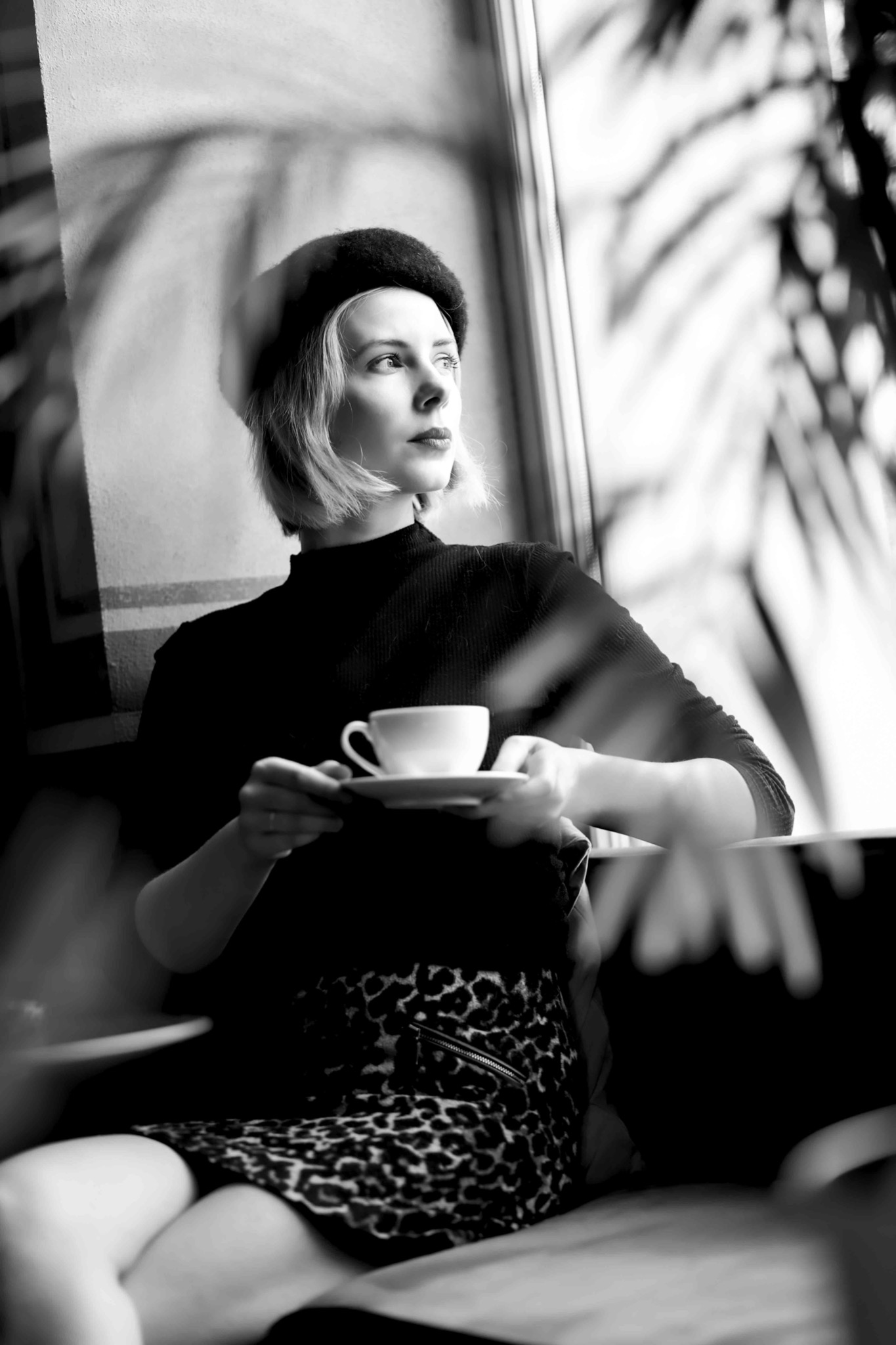 Czarno-białe zdjęcie kobiety w berecie, trzymającej filiżankę kawy, siedzącej przy oknie z widocznymi cieniami roślin