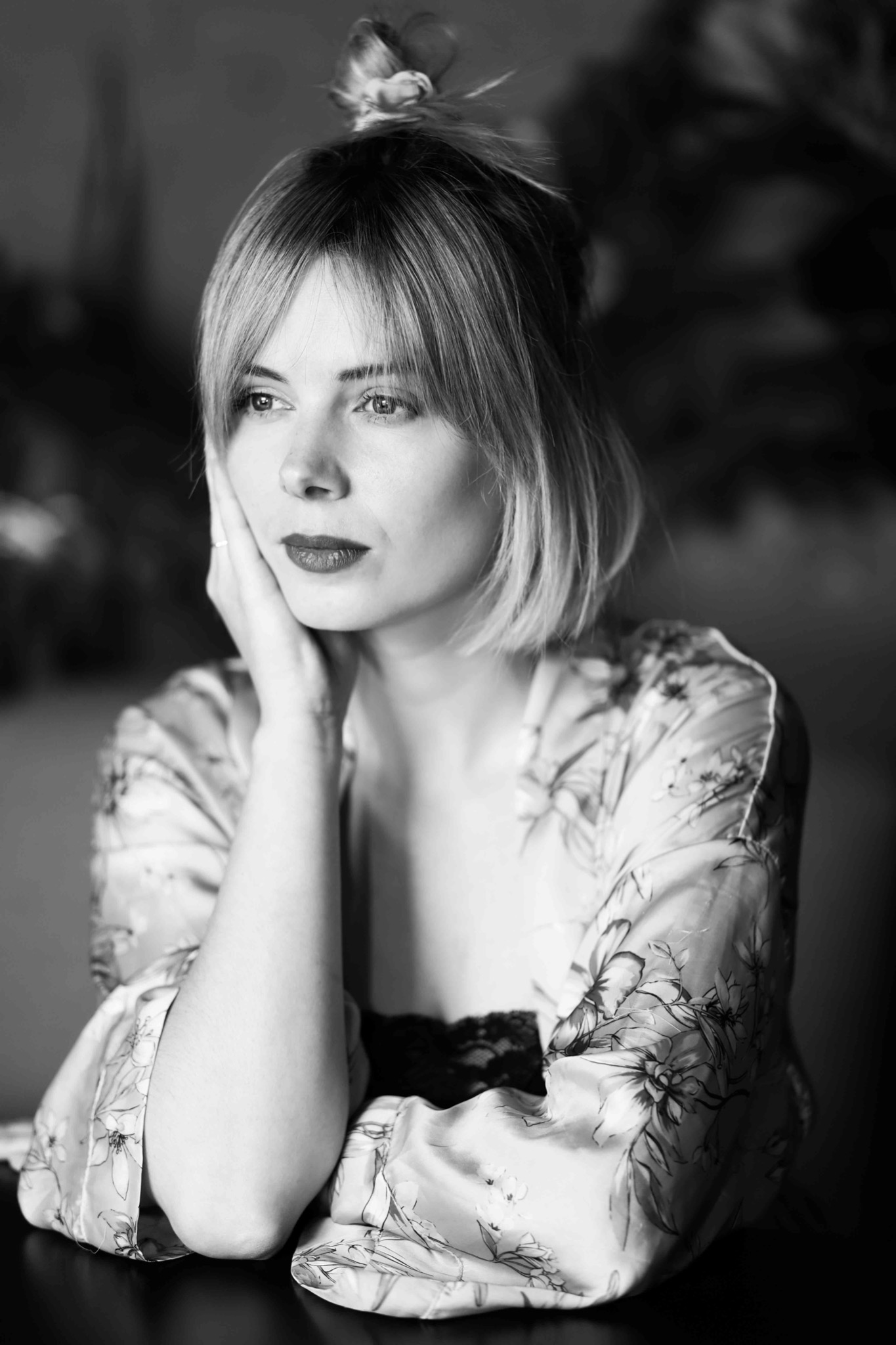 Portret kobiety z blond grzywką, w kwiecistym szlafroku, opierającej twarz na dłoni, czarno-biały kadr.