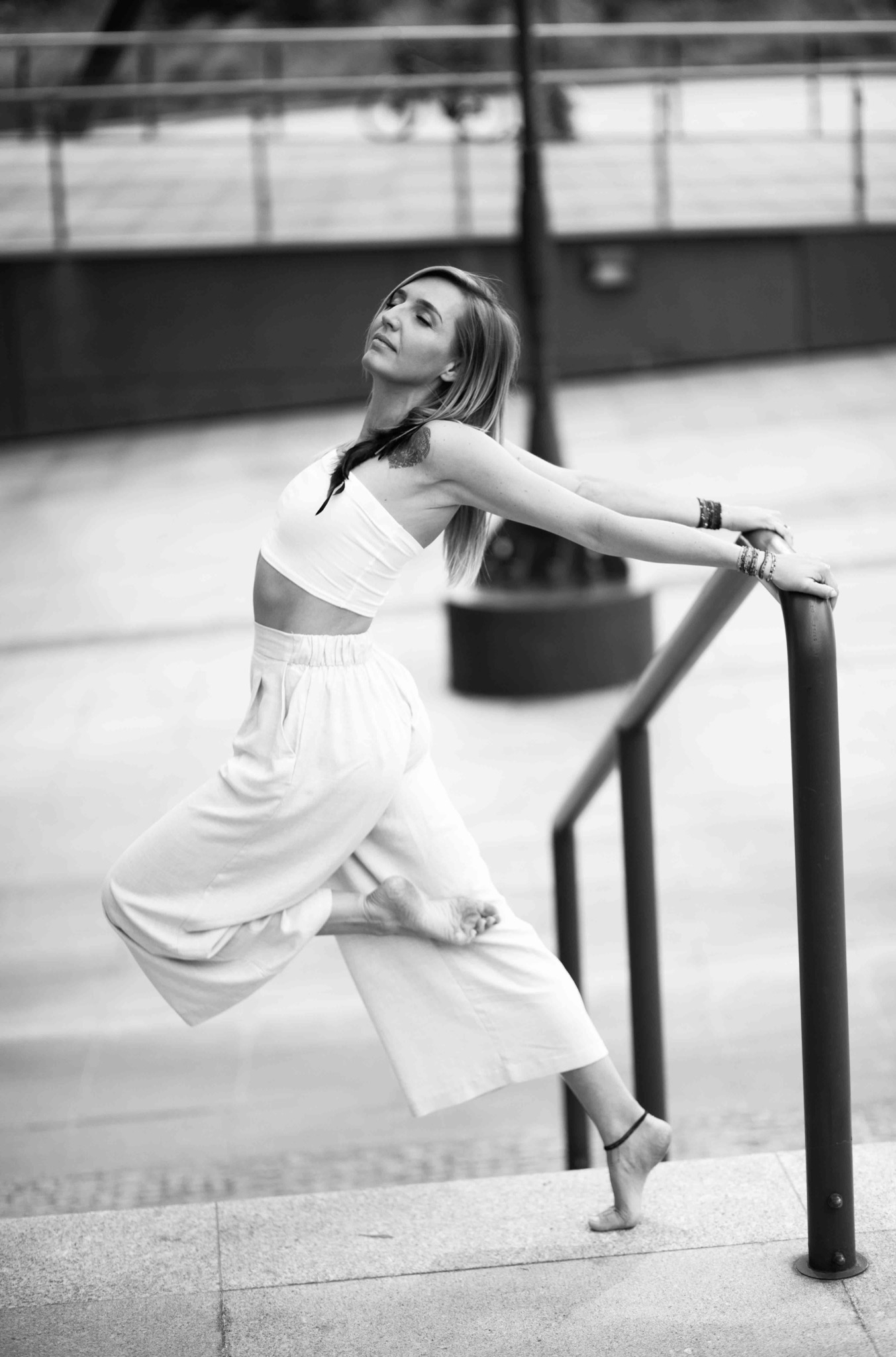 Czarno-białe zdjęcie kobiety w białym stroju, opierającej się o poręcz, z zamkniętymi oczami i tatuażem na ramieniu.