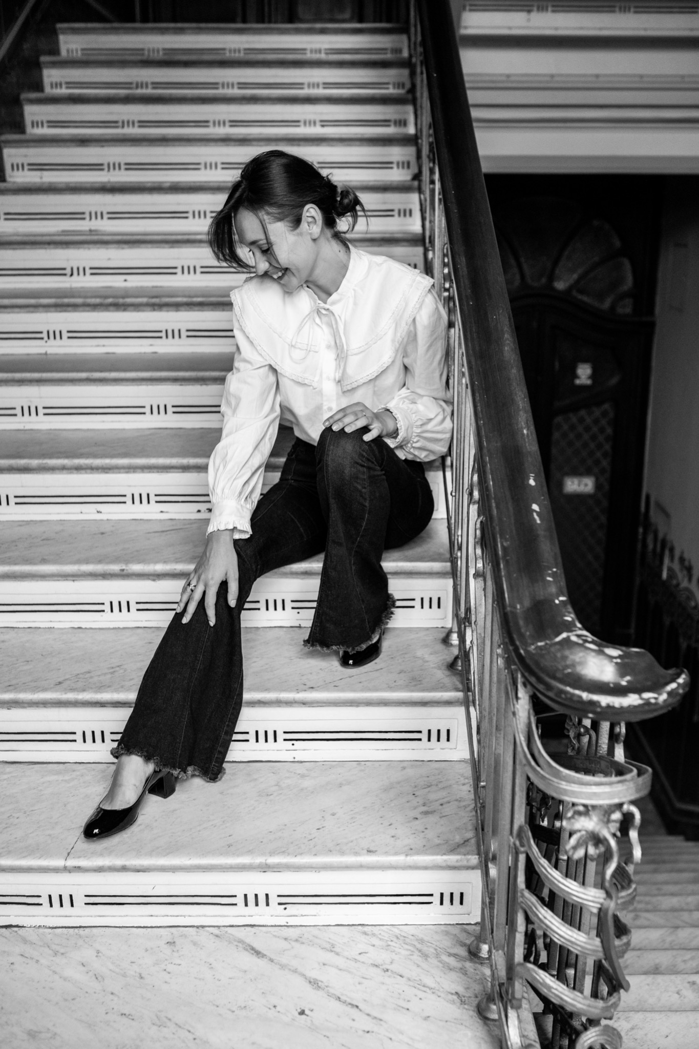 Kobieta w białej bluzce i dżinsach siedzi na marmurowych schodach z metalową balustradą, spoglądając w dół z uśmiechem.