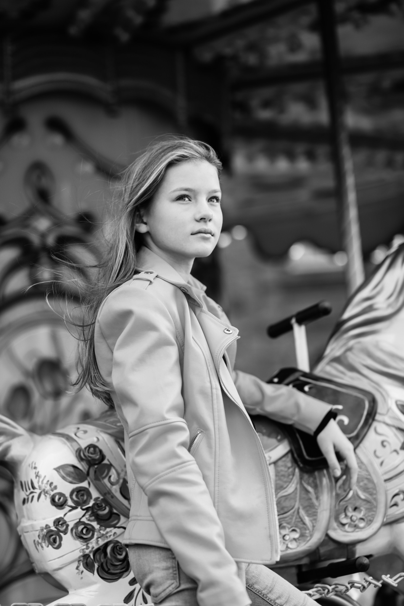 Czarno-białe zdjęcie nastolatki w jasnej kurtce, siedzącej na karuzelowym koniu z ręcznie malowanymi kwiatami, zamyślona poza i wiatr we włosach.