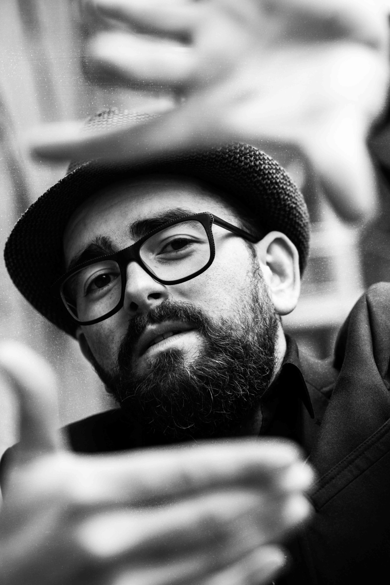 Portret mężczyzny z brodą i okularami w kapeluszu, czarno-biały, z ręką w kadrze, skierowany w stronę obiektywu.