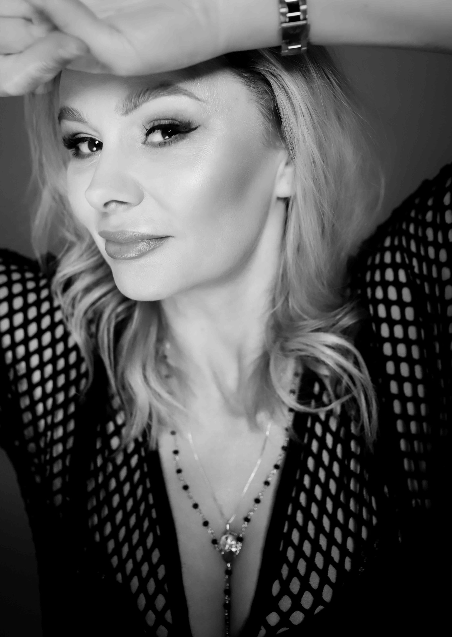Portret kobiety w czarno-białej tonacji, z delikatnym uśmiechem, w bluzce z siateczki i naszyjnikiem, dłoń z zegarkiem nad czołem