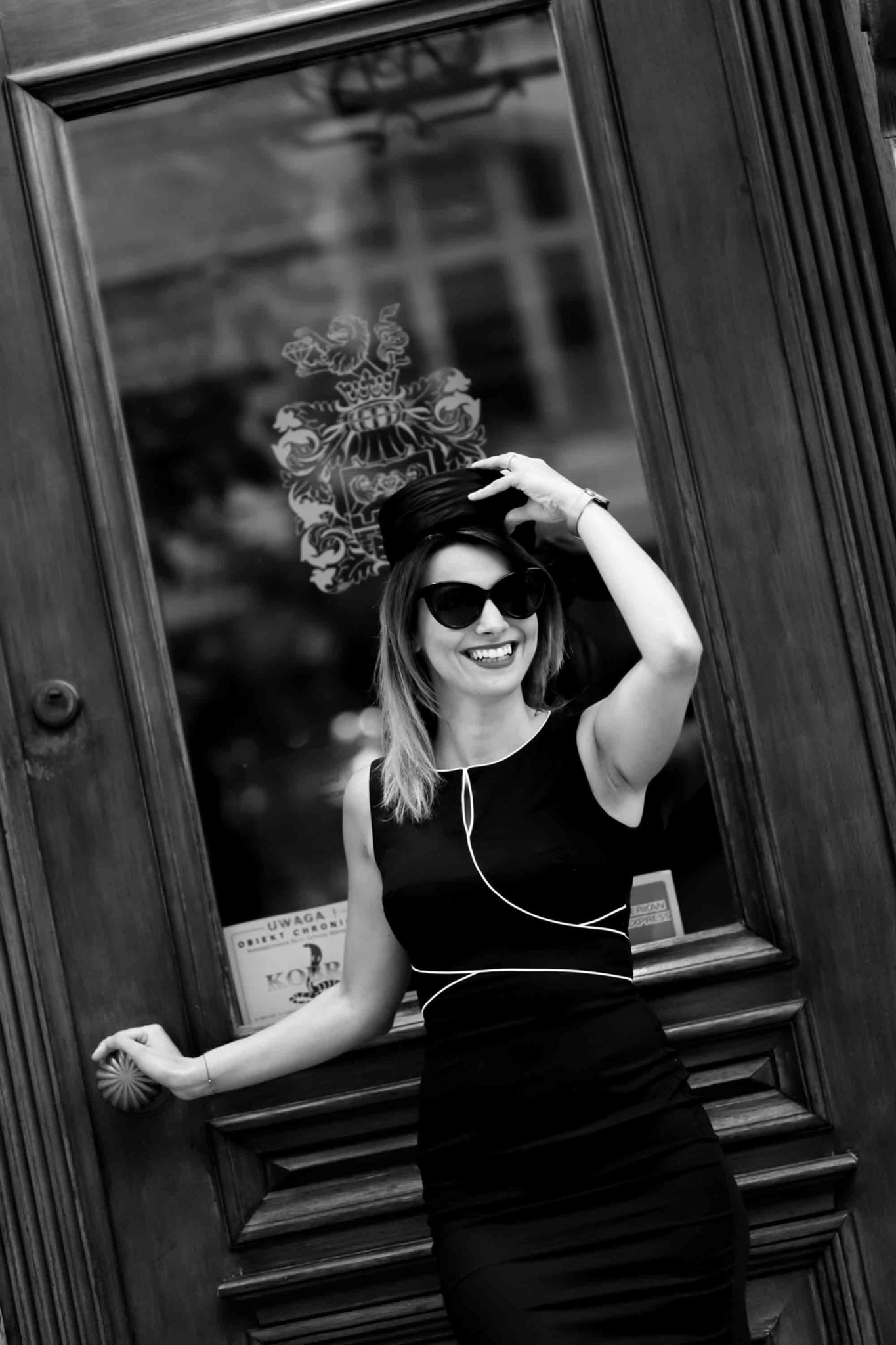 Uśmiechnięta kobieta w czarnej sukience i kapeluszu, z okularami przeciwsłonecznymi, opiera się o ciemne, drewniane drzwi z dekoracyjnym, wytrawionym wzorem na szkle i tabliczką informacyjną