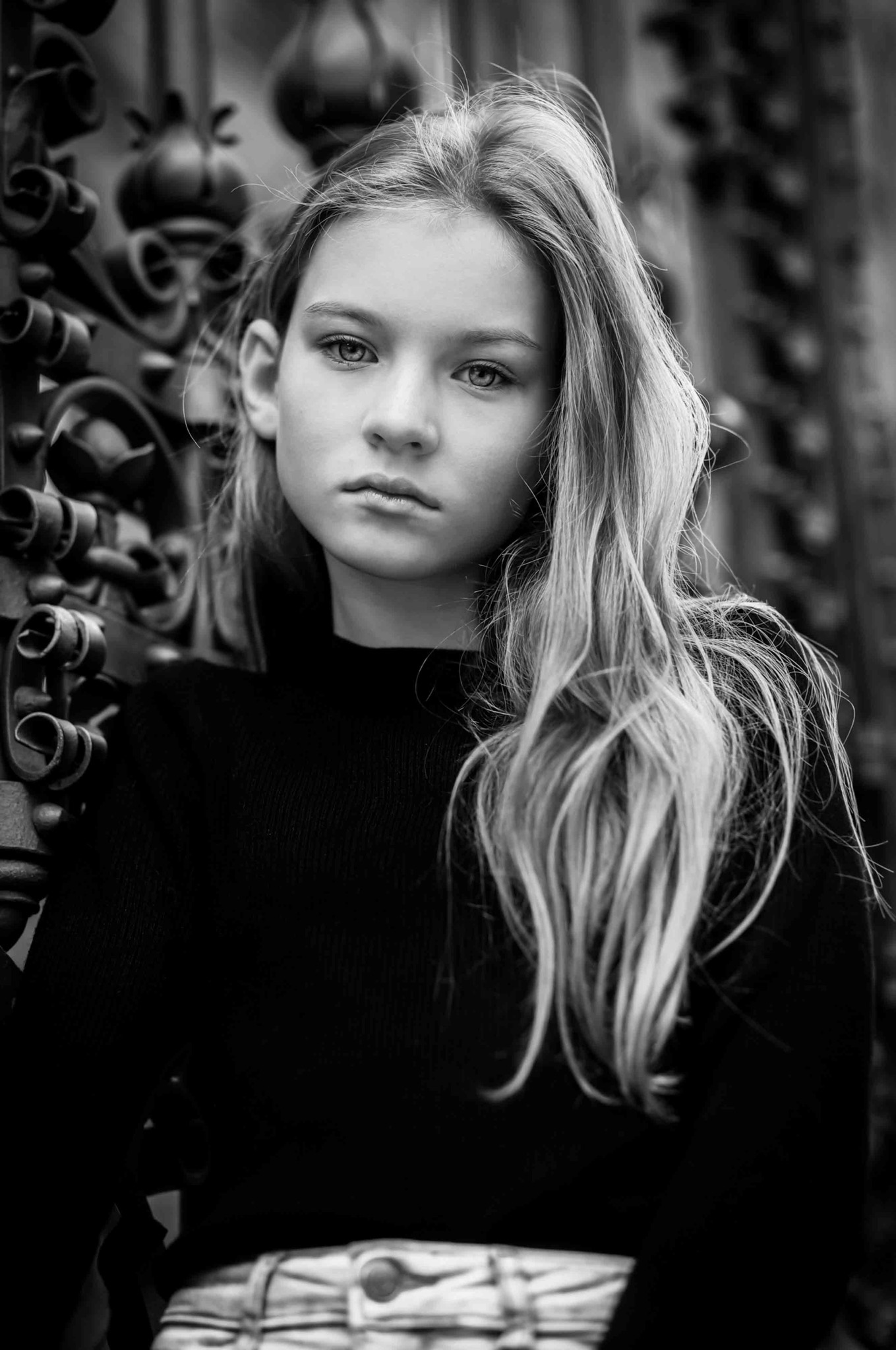 Portret młodej kobiety o jasnych włosach, opierającej się o ozdobne, żelazne ogrodzenie, w czarno-białej tonacji.