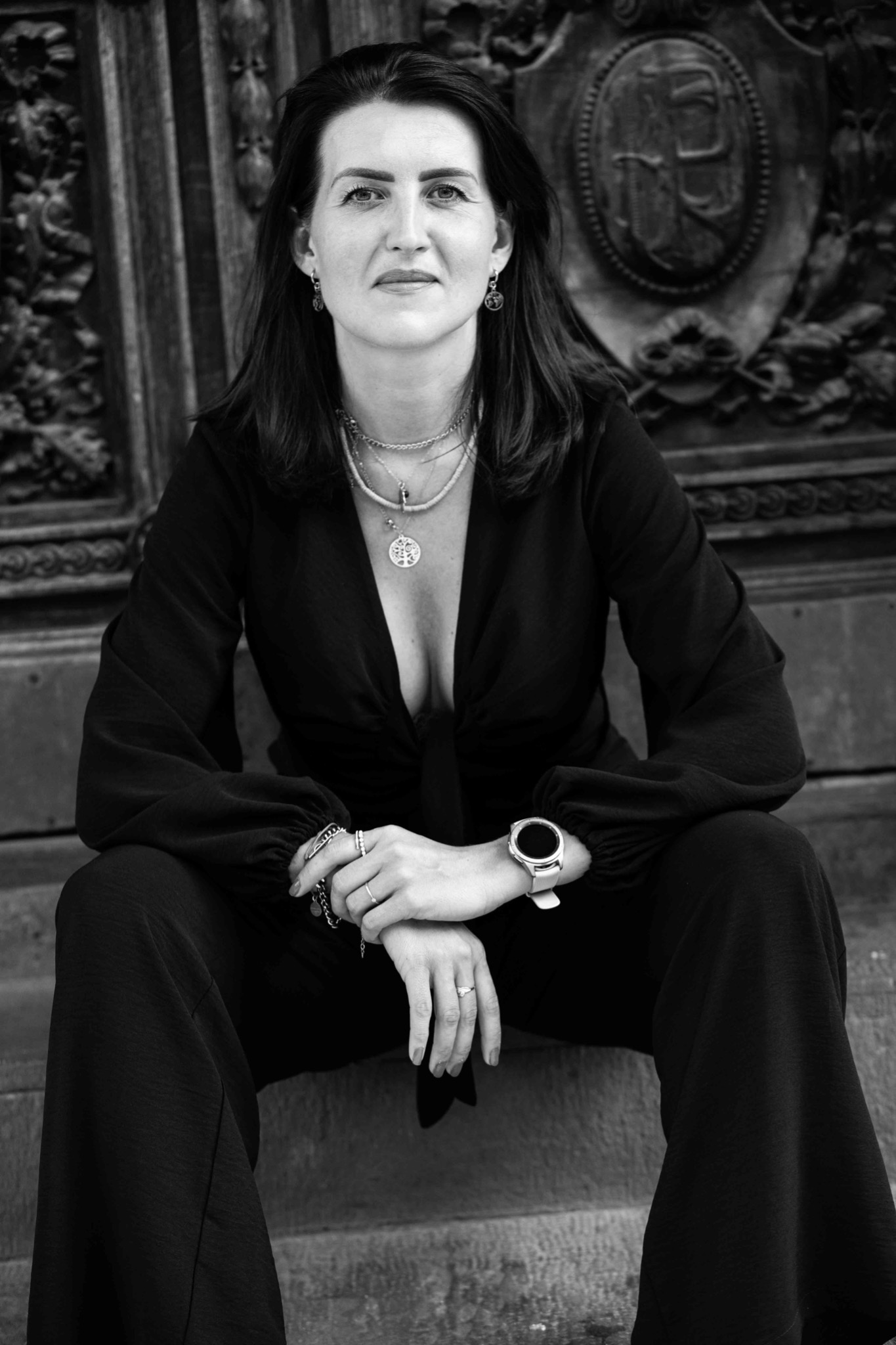 Portret kobiety w czarnym kombinezonie siedzącej na kamiennych schodach na tle zdobionych, drewnianych drzwi, z widoczną biżuterią i zegarkiem na ręce, w czarno-białej tonacji.