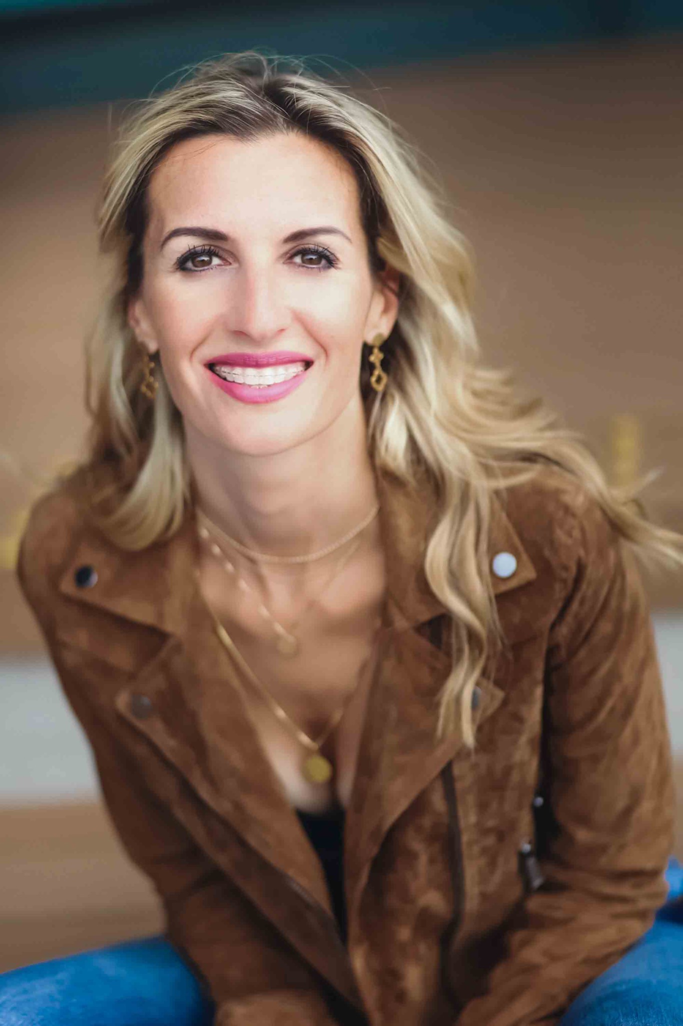 Uśmiechnięta blondynka w brązowej zamszowej kurtce, z aparatem ortodontycznym, złotymi kolczykami i naszyjnikiem, siedząca na tle brązowej ściany.