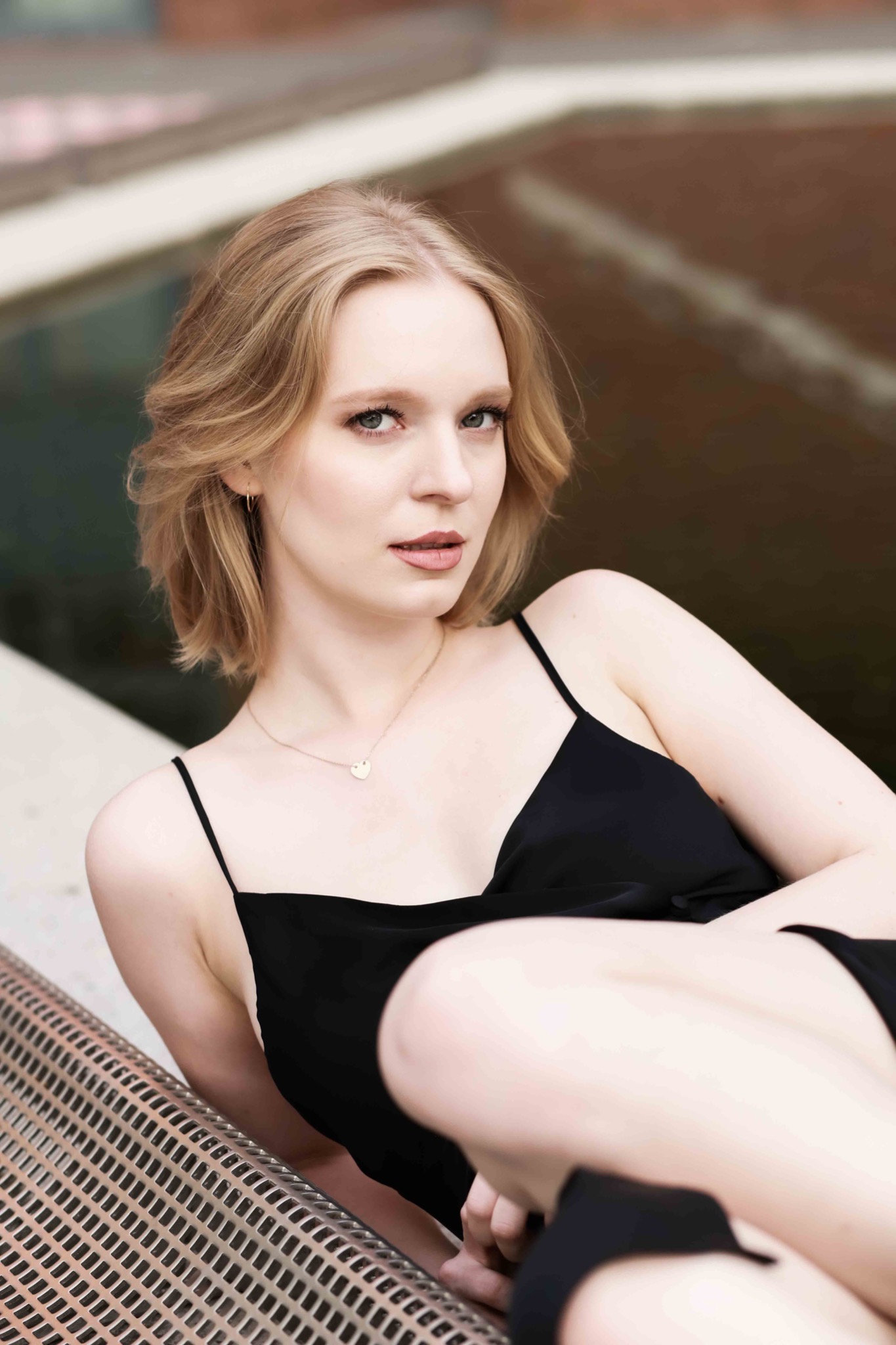 Portret kobiety o jasnych włosach w czarnej sukience, leżącej na metalowej ławce z tłem w odcieniach brązu.