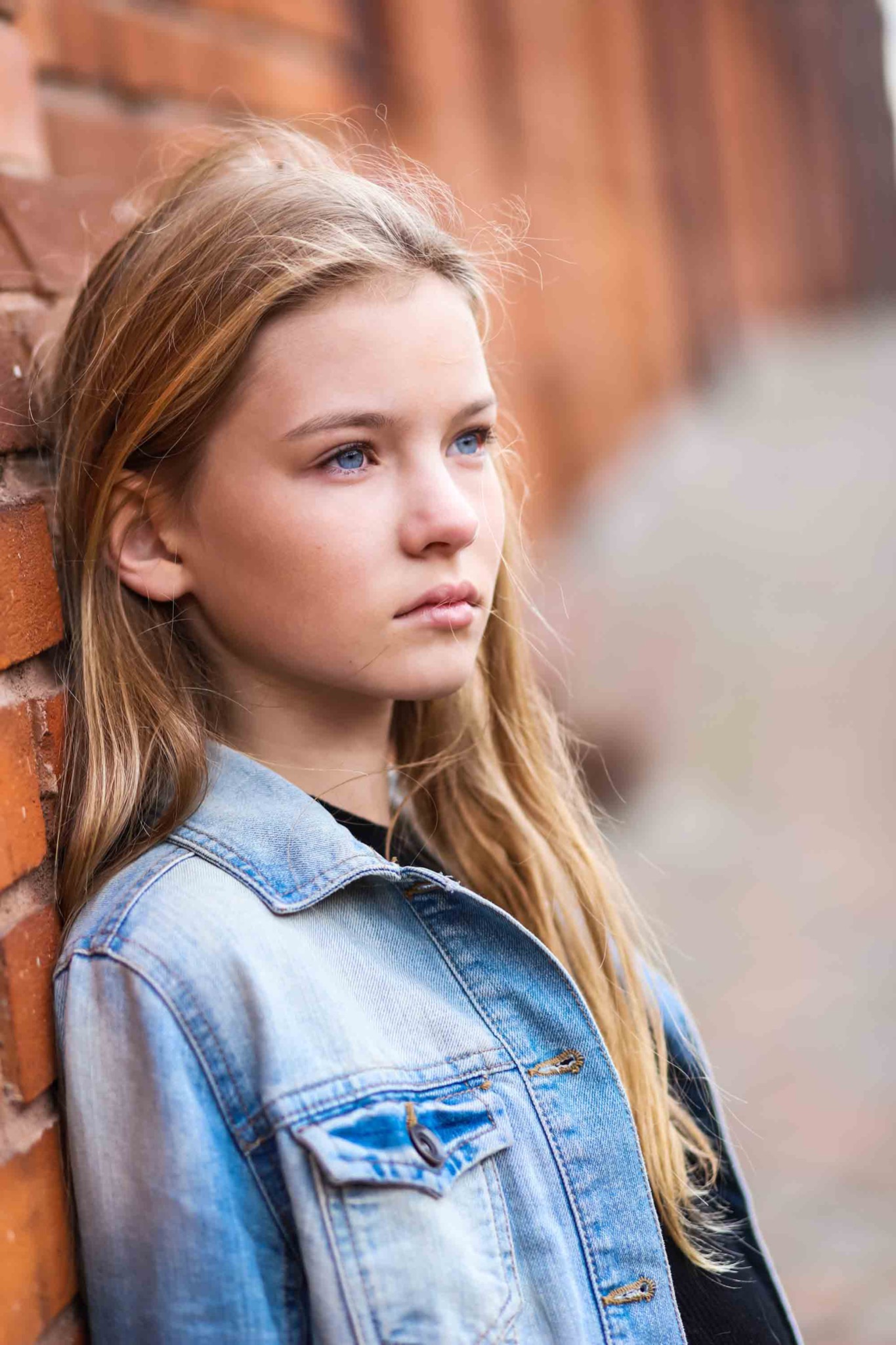 Portret nastolatki z blond włosami i niebieskimi oczami, oparta o ceglaną ścianę, ubrana w dżinsową kurtkę, zamyślona poza, naturalne światło.