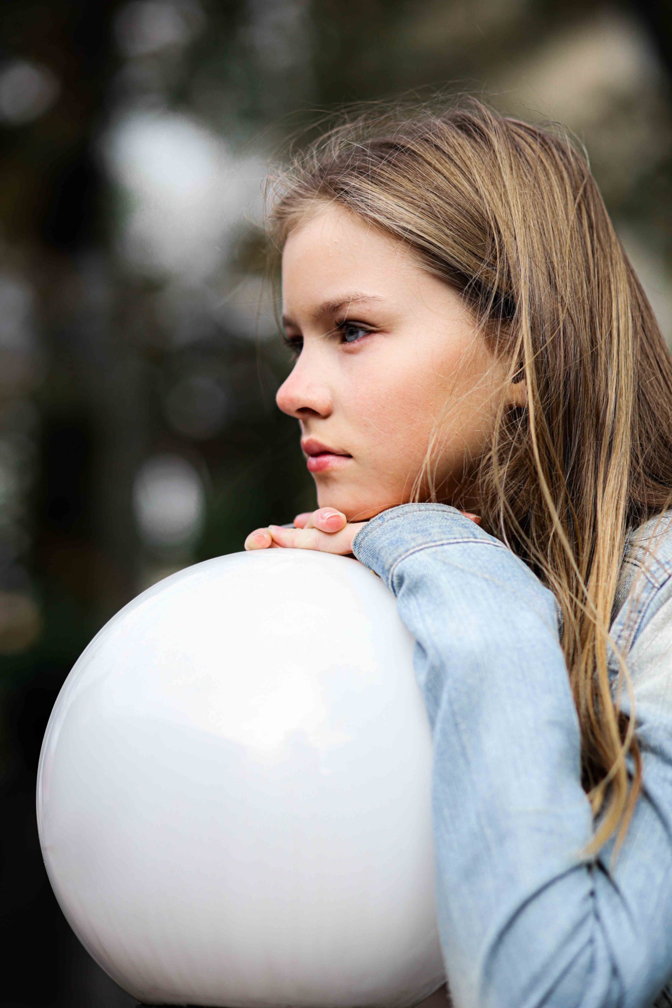 Portret z profilu młodej dziewczyny z blond włosami opierającej podbródek na białej, okrągłej lampie w plenerze.