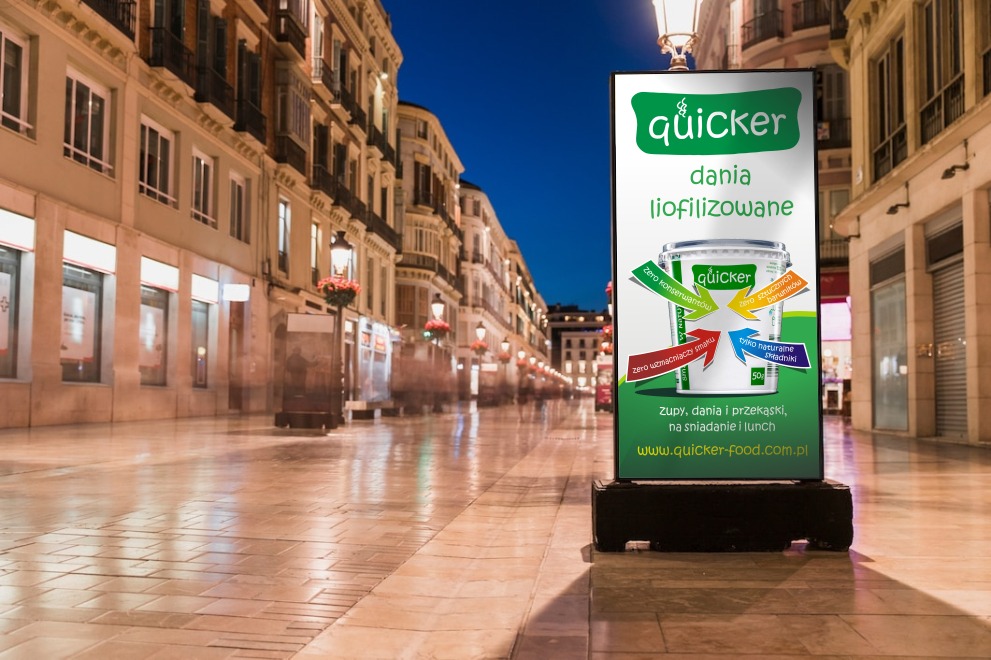 Billboard reklamowy z logo Quicker i hasłem 'dania liofilizowane' ustawiony na pustej ulicy miejskiej o zmierzchu.