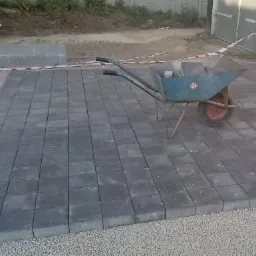 Fragment układanej nawierzchni z szarych kostek brukowych, widoczny niebieski wózek z kostką i biało-czerwona taśma ostrzegawcza.