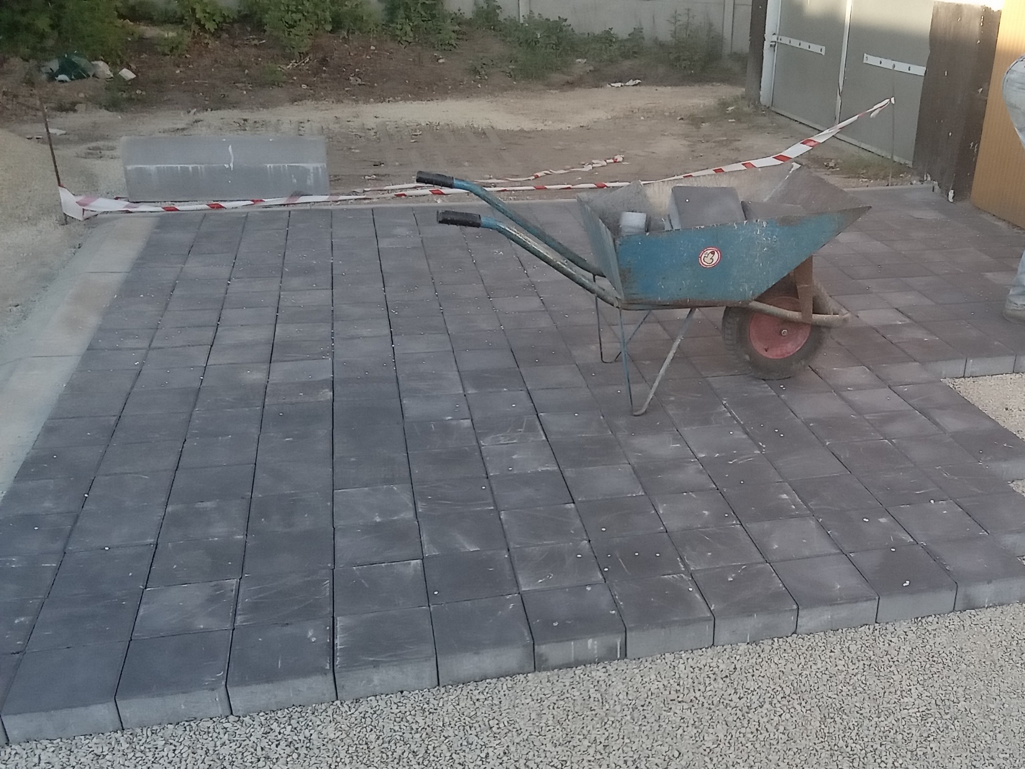 Fragment układanej nawierzchni z szarych kostek brukowych, widoczny niebieski wózek z kostką i biało-czerwona taśma ostrzegawcza.