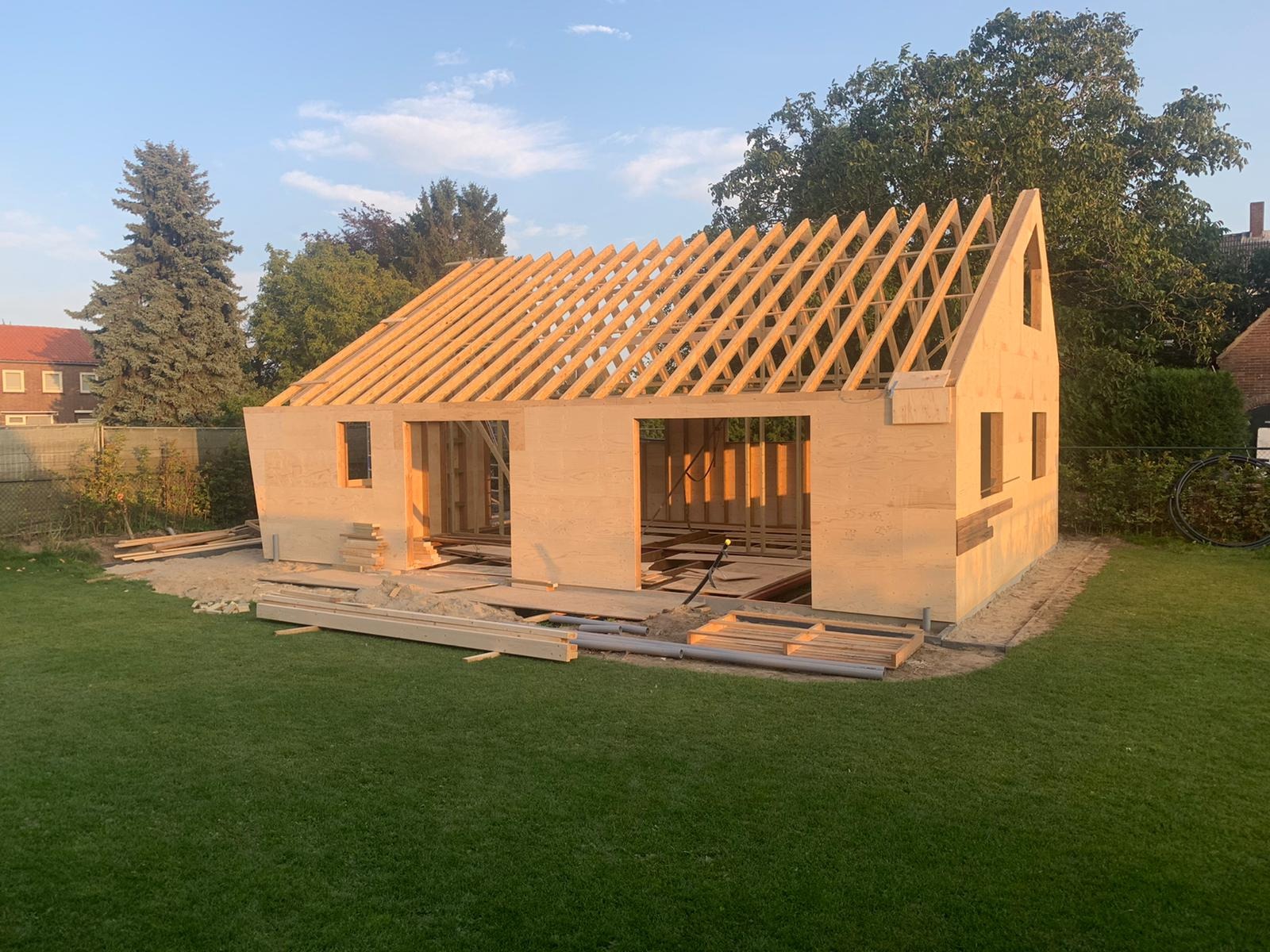 Konstrukcja drewnianego budynku w trakcie budowy, widoczne ściany z otworami na okna i drzwi oraz szkielet dachu z łat, na trawniku leżą materiały budowlane.