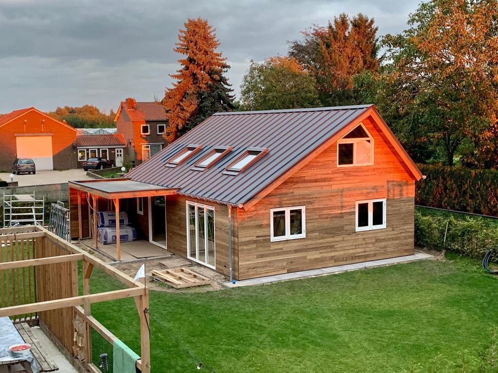 Nowoczesny dom z drewnianą elewacją i metalowym dachem, widok z góry na ogród i okolicę, wczesny etap budowy z widocznymi materiałami budowlanymi na tarasie.