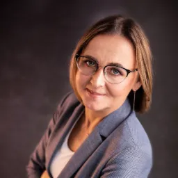 Portret kobiety w okularach i szarym żakiecie na ciemnym tle, uśmiechająca się, z delikatnym makijażem.