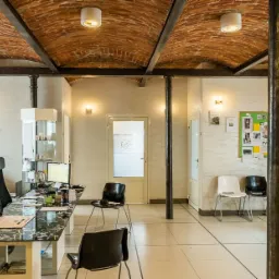 Wnętrze poczekalni z recepcją: biurko z czarnym blatem i krzesłami, ściany w jasnym odcieniu, sklepienie z cegły, oświetlenie punktowe i kolumny.