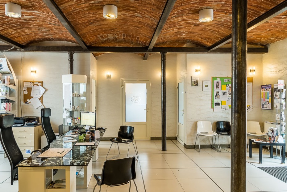 Wnętrze poczekalni z recepcją: biurko z czarnym blatem i krzesłami, ściany w jasnym odcieniu, sklepienie z cegły, oświetlenie punktowe i kolumny.