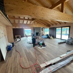 Montaż drewnianej podłogi w przestronnym, jasnym pomieszczeniu z drewnianym stropem, widoczne narzędzia i materiały budowlane, zarys sylwetki pracownika na kolanach.