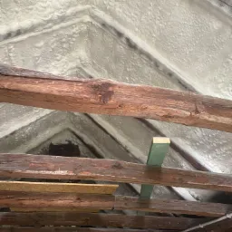 Poddasze ocieplone pianką PUR, widoczne drewniane belki konstrukcyjne, częściowo zakryte pianą, surowe drewno w dolnej części kadru i fragment instalacji.