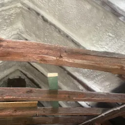 Poddasze z widocznymi drewnianymi belkami konstrukcyjnymi, pokryte grubą warstwą białej pianki izolacyjnej, z fragmentem zielonej deski pomiędzy belkami.