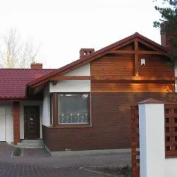 Dwukondygnacyjny dom jednorodzinny z garażem, z czerwonym dachem i ceglaną elewacją, częściowo wykończony drewnem. Widoczny fragment ogrodzenia.