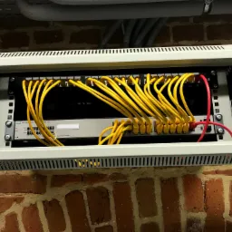 Panel krosowniczy z żółtymi kablami sieciowymi podłączonymi do przełącznika HPE OfficeConnect 1920S, zamontowany na ścianie z czerwonej cegły.