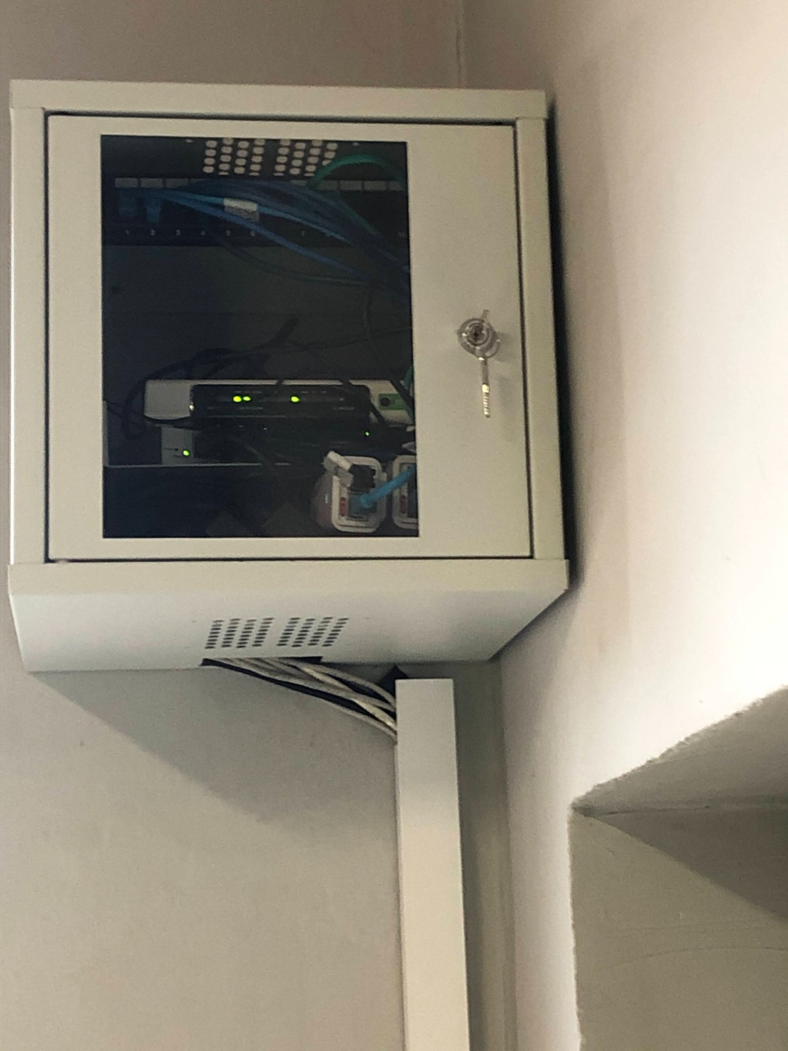 Ścienna szafa serwerowa z zamkiem, zawierająca router i przełącznik sieciowy z podłączonymi kablami Ethernet, umieszczona w rogu pomieszczenia.