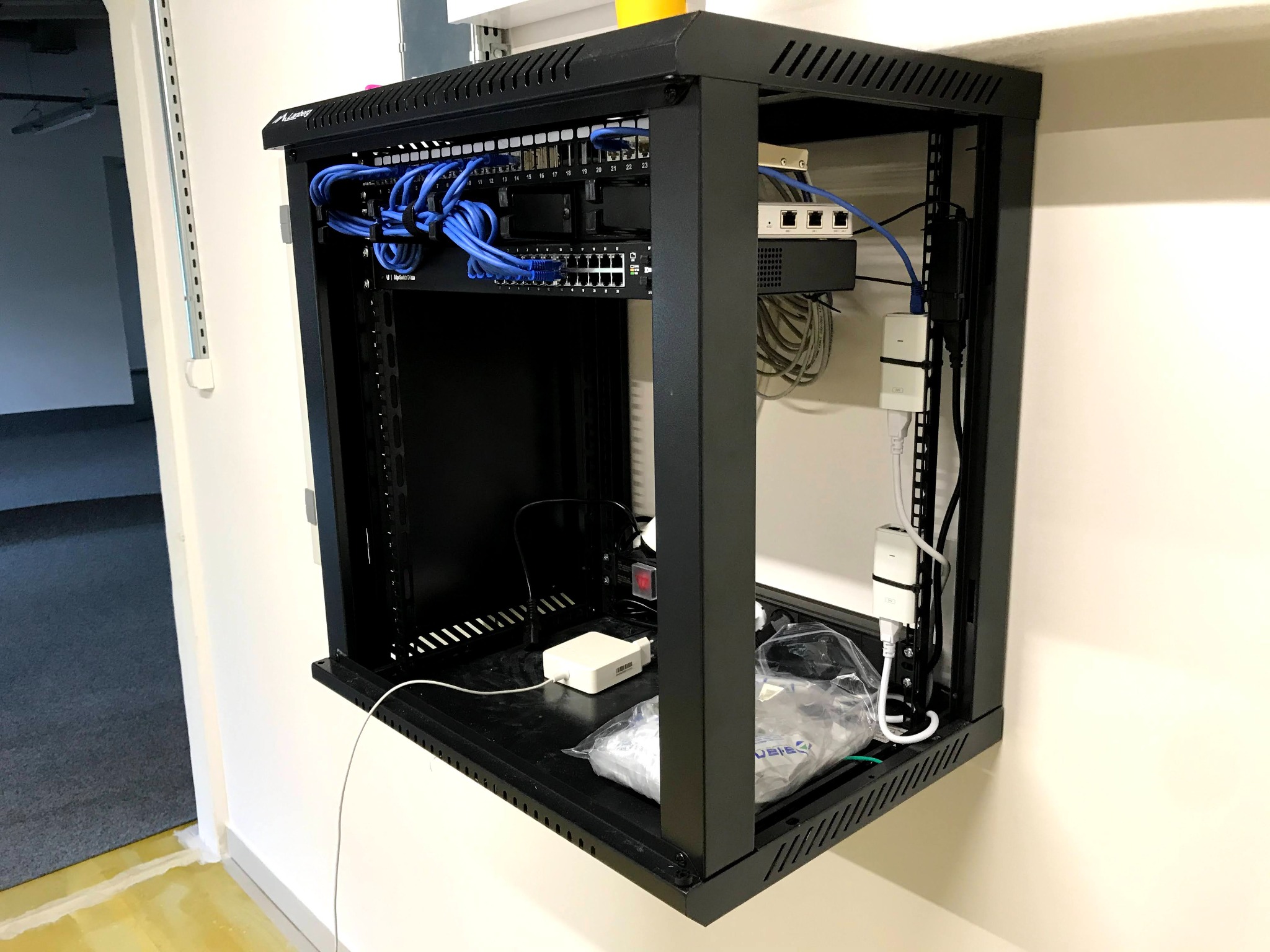 Ściana z zawieszoną szafą teleinformatyczną z patch panelem, switchem i routerem, okablowanie sieciowe w kolorze niebieskim, dodatkowe zasilacze i kable wewnątrz szafy, widok z boku na białą ścianę...