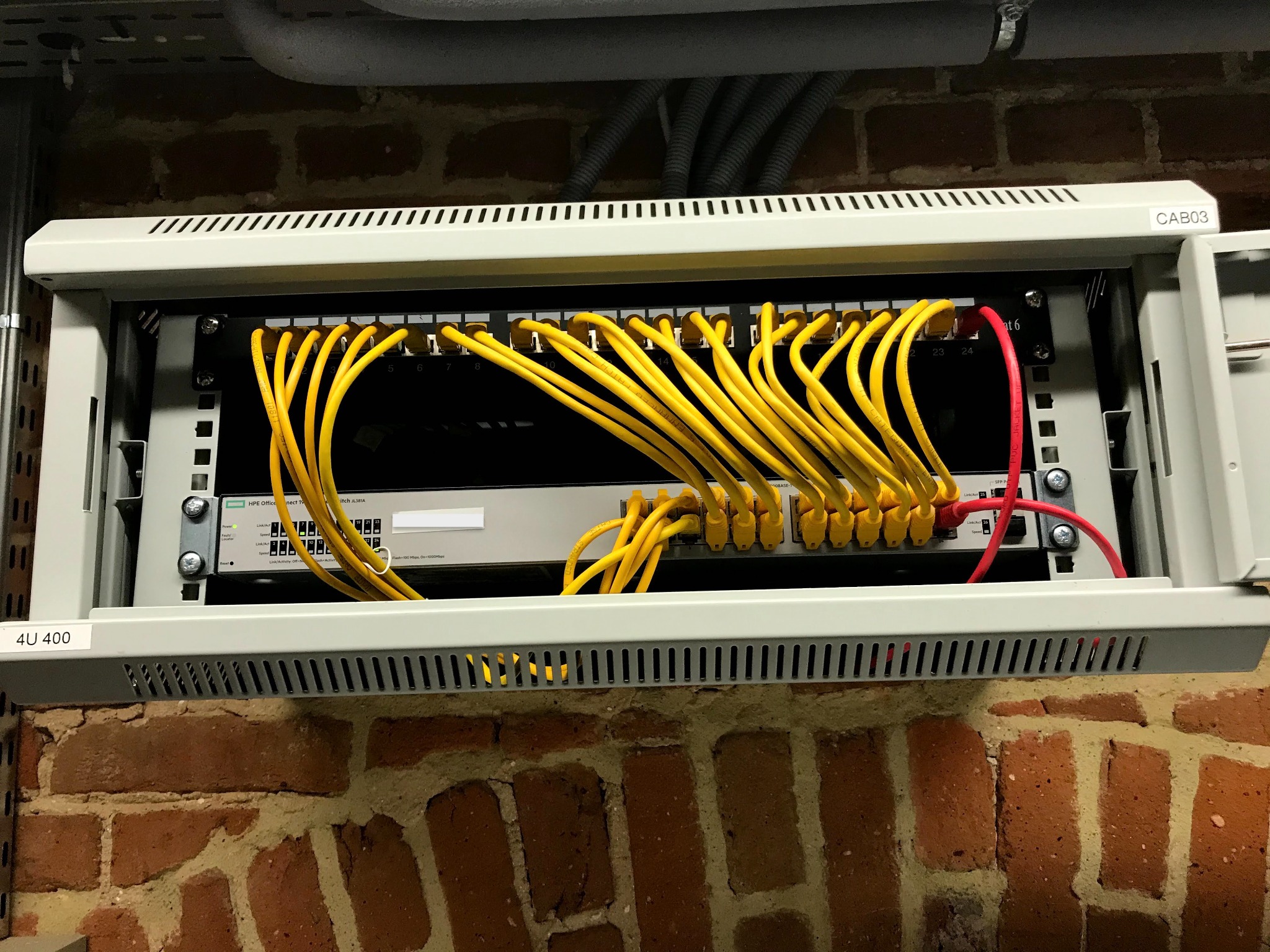 Panel krosowniczy z żółtymi kablami sieciowymi podłączonymi do przełącznika HPE OfficeConnect 1920S, zamontowany na ścianie z czerwonej cegły.