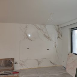KP Remonty Kamil Pierzakowski - Ściana wyłożona płytami imitującymi marmur z przygotowanym miejscem na telewizor, widoczne kable od oświetlenia sufitowego, folia ochronna na podłodze, okno z czarną ramą.