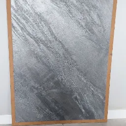 Próbka dekoracyjnej, srebrnej farby strukturalnej na płycie MDF, prezentująca efekt przecierki, ustawiona na szarych płytkach podłogowych przy białej ścianie.