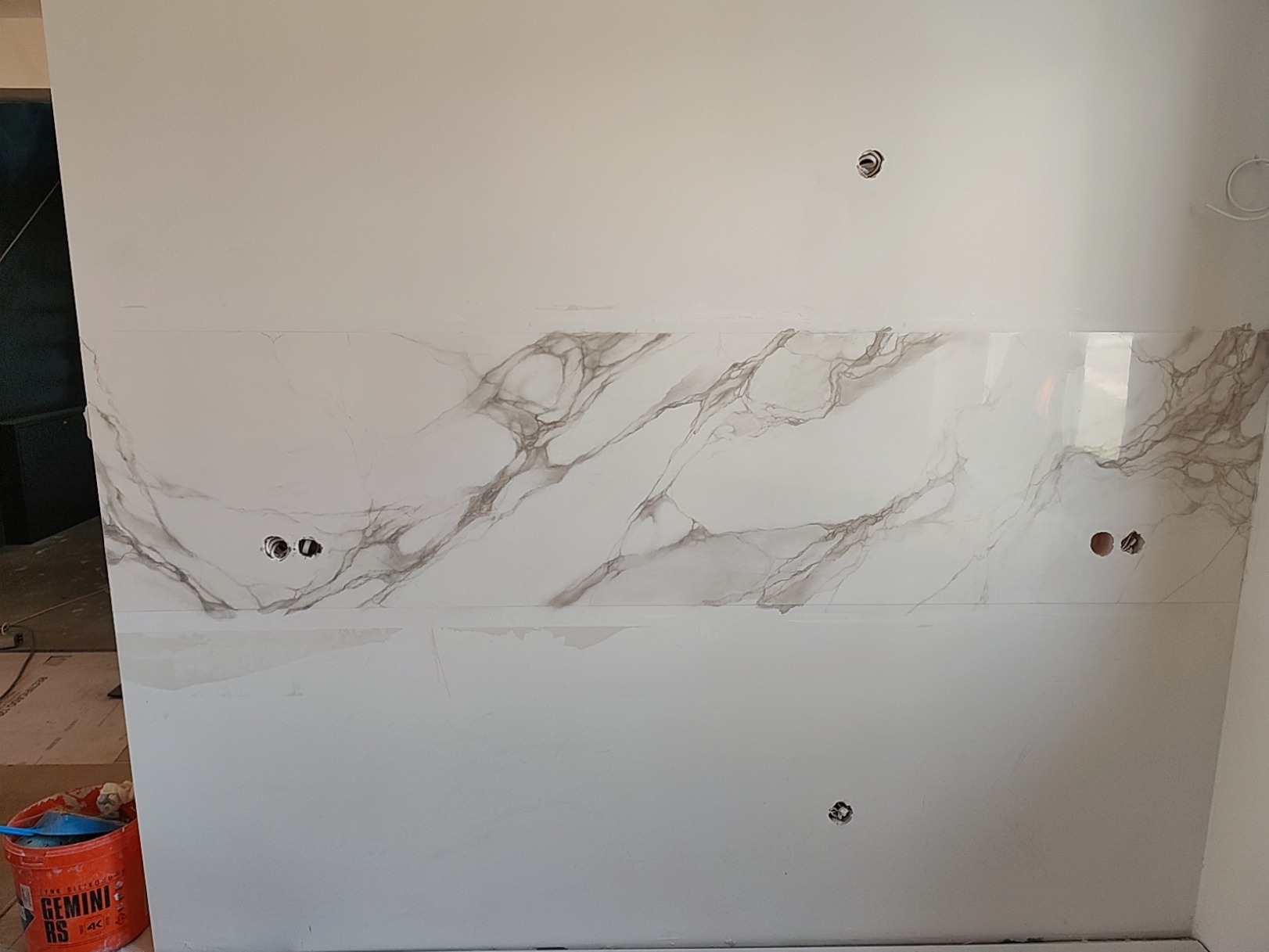 Ściana z częściowo położonymi płytami imitującymi marmur, widoczne puszki elektryczne, poniżej przygotowana powierzchnia do dalszych prac.