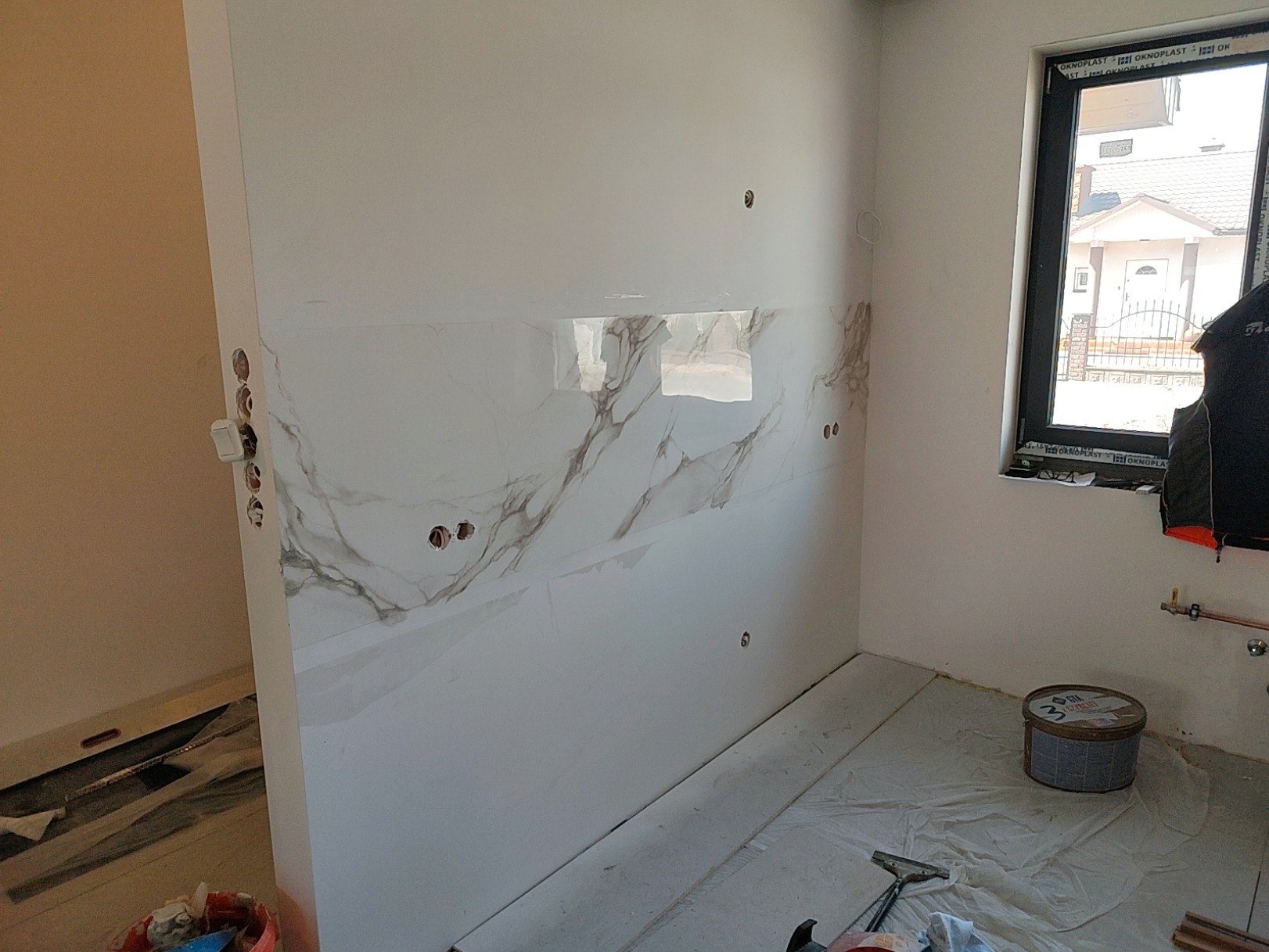 Ściana wyłożona białymi płytkami imitującymi marmur w trakcie prac wykończeniowych, widoczne puszki elektryczne, okno z widokiem na dom, wiadro z farbą i folia ochronna na podłodze.