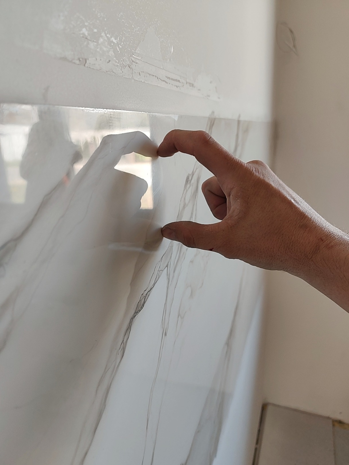Ręka mierzy odstęp między płytkami imitującymi marmur na ścianie, widoczne ślady po szpachlowaniu nad płytkami.
