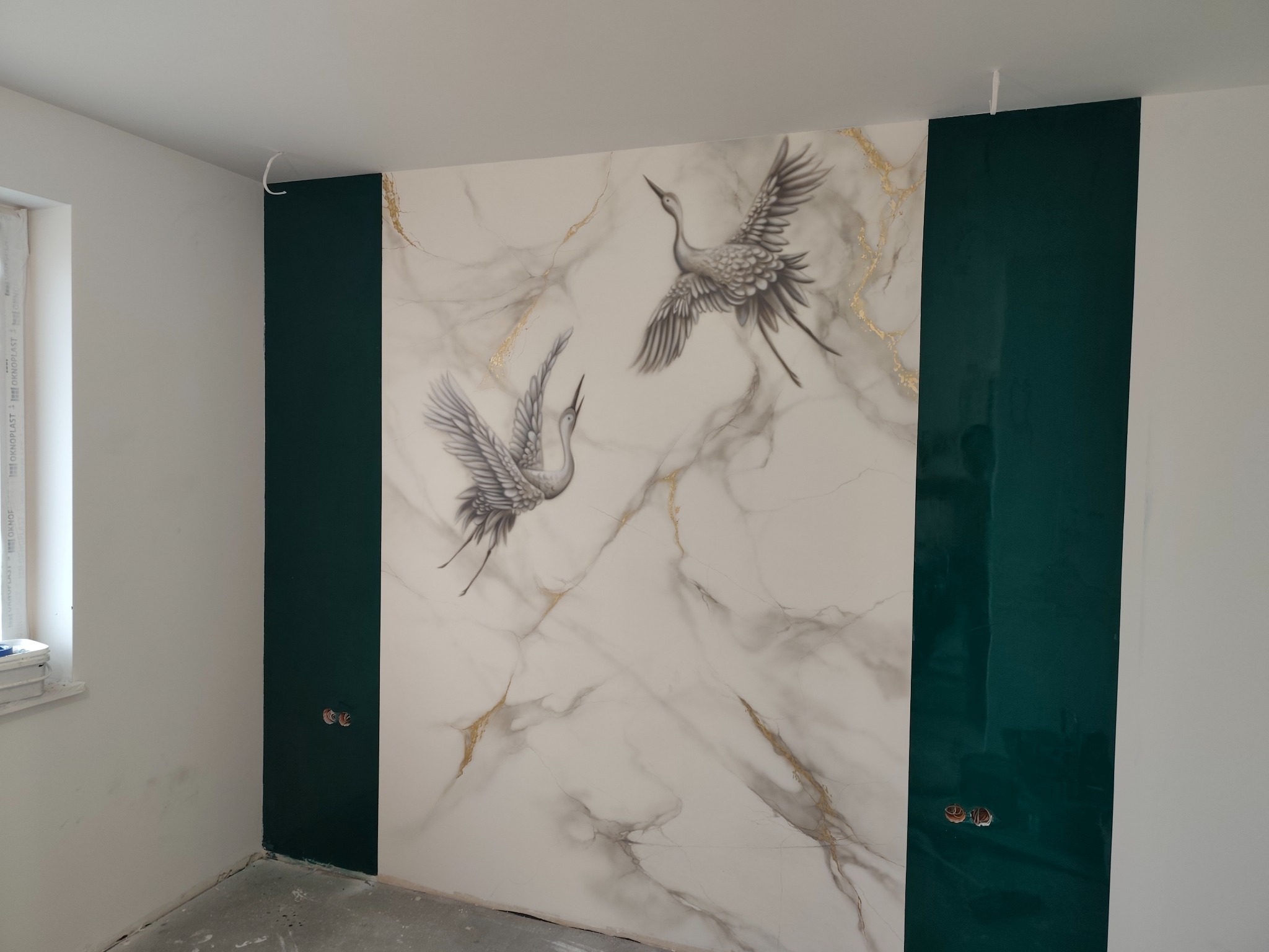 Ściana w Dobiegniewie z dekoracyjnym malowaniem imitującym marmur z motywem żurawi w locie, obok pionowe pasy w kolorze butelkowej zieleni, widoczne gniazdka elektryczne.