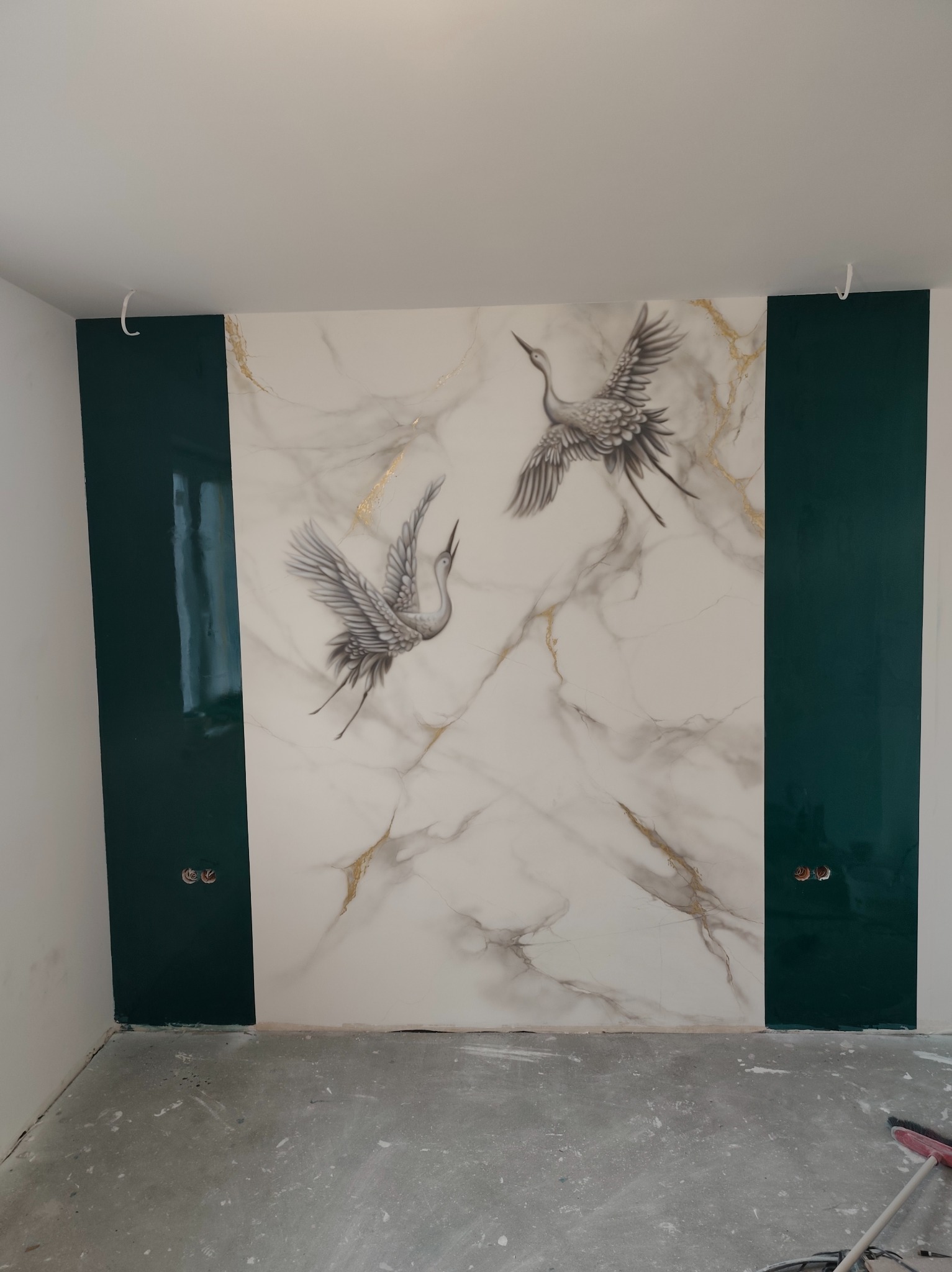 Ściana z marmurowym wzorem i motywem żurawi, obramowana ciemnozielonymi panelami, widoczne gniazdka elektryczne i surowa podłoga w trakcie wykańczania.