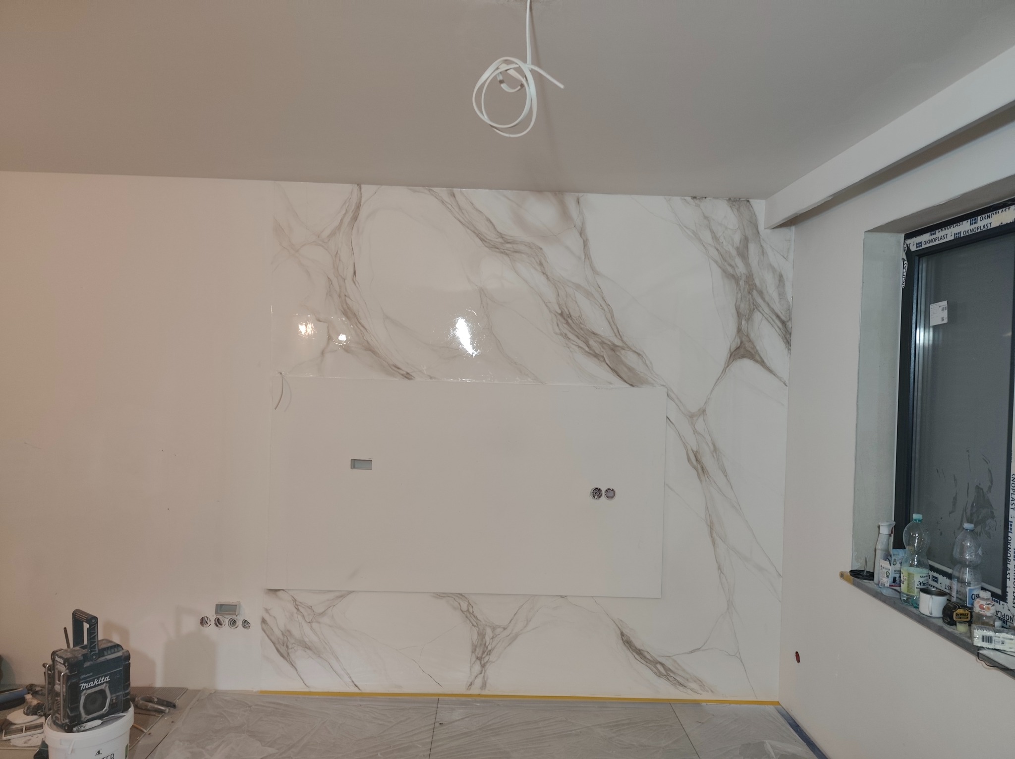 Ściana wykończona płytami imitującymi marmur z białym panelem centralnym i przygotowanymi otworami na gniazdka, widoczne narzędzia i sprzęt budowlany, folia ochronna na podłodze, surowe przewody...