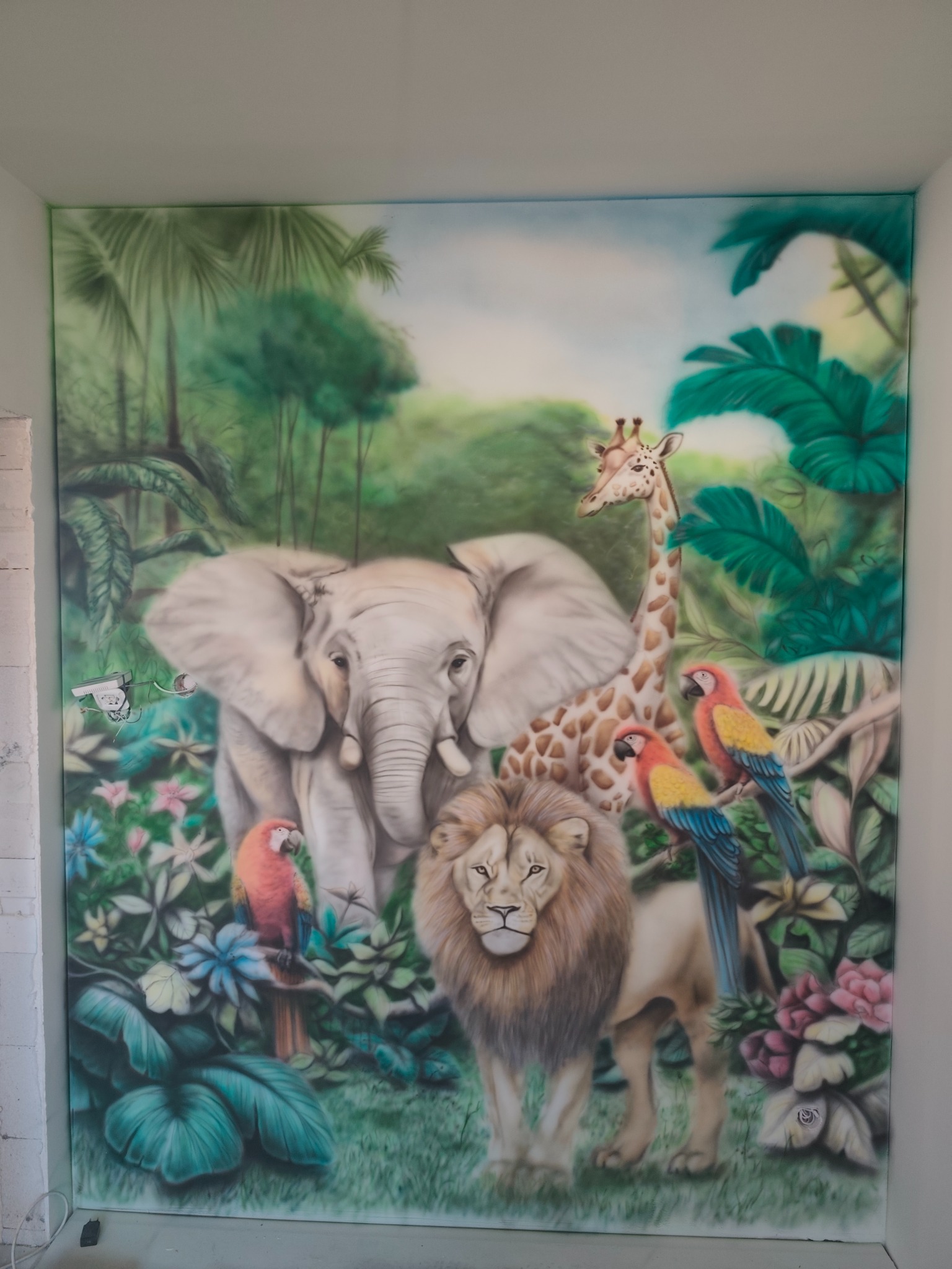 Ściana ozdobiona malowidłem przedstawiającym afrykańską dżunglę z lwem, słoniem, żyrafą i papugami, wykonanym techniką aerografu.