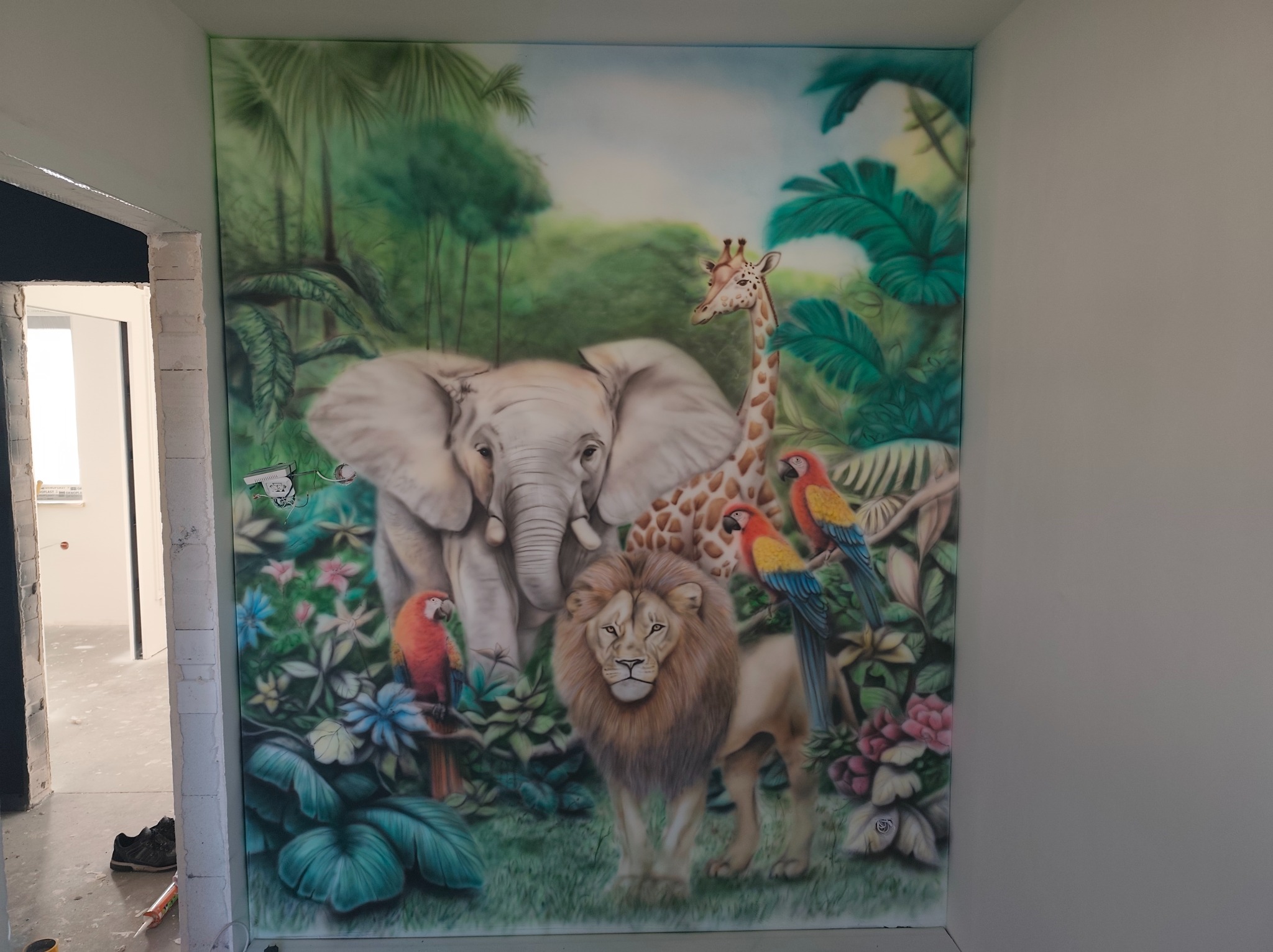 Malowidło ścienne w pokoju dziecięcym przedstawiające afrykańską dżunglę z lwem, słoniem, żyrafą i papugami, widoczne obok surowego muru z cegieł i niedokończonego przejścia.