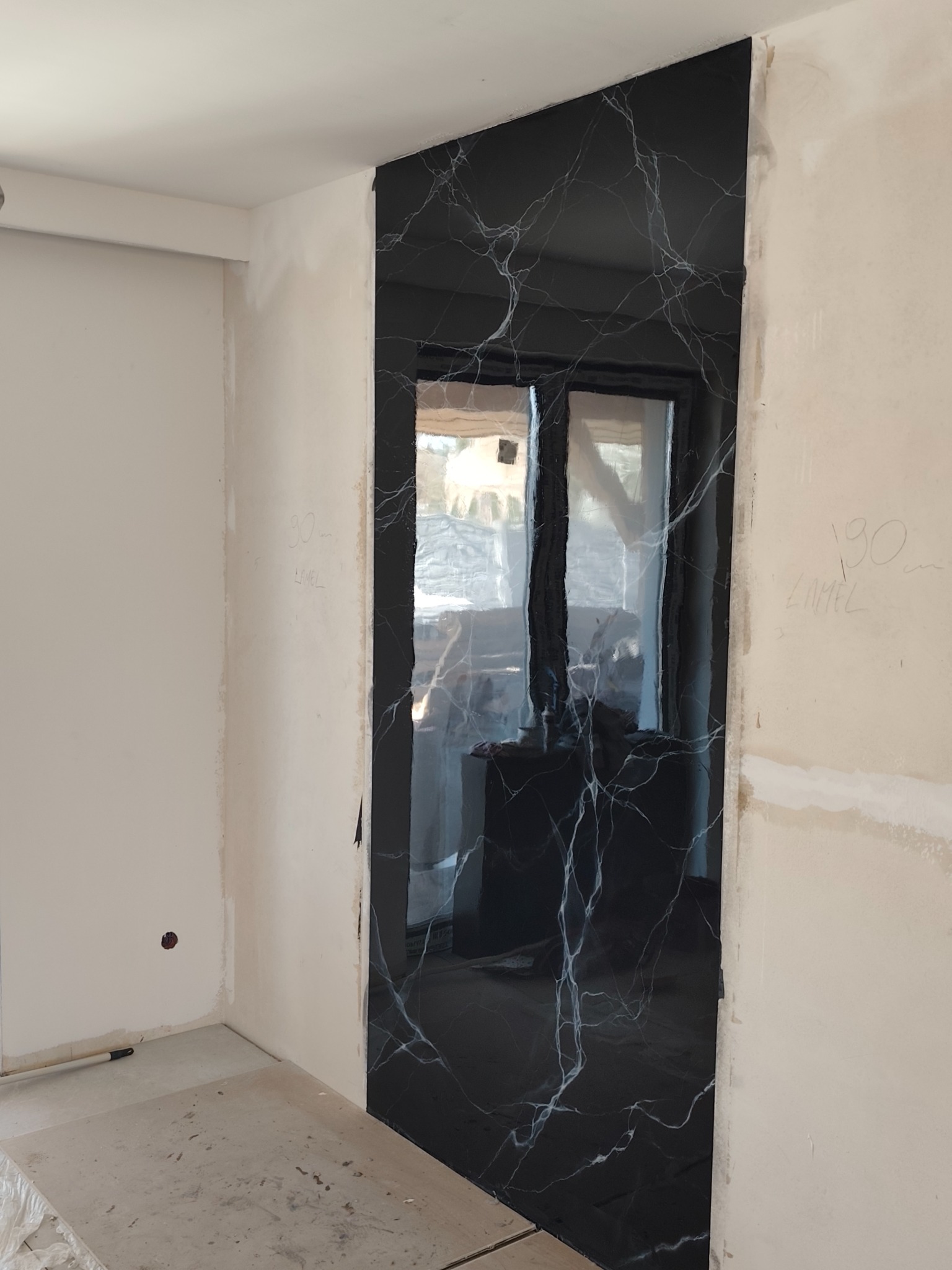 Fragment ściany z czarną płytą imitującą marmur z białymi żyłkami, w trakcie prac wykończeniowych, widoczne surowe ściany i podłoga.