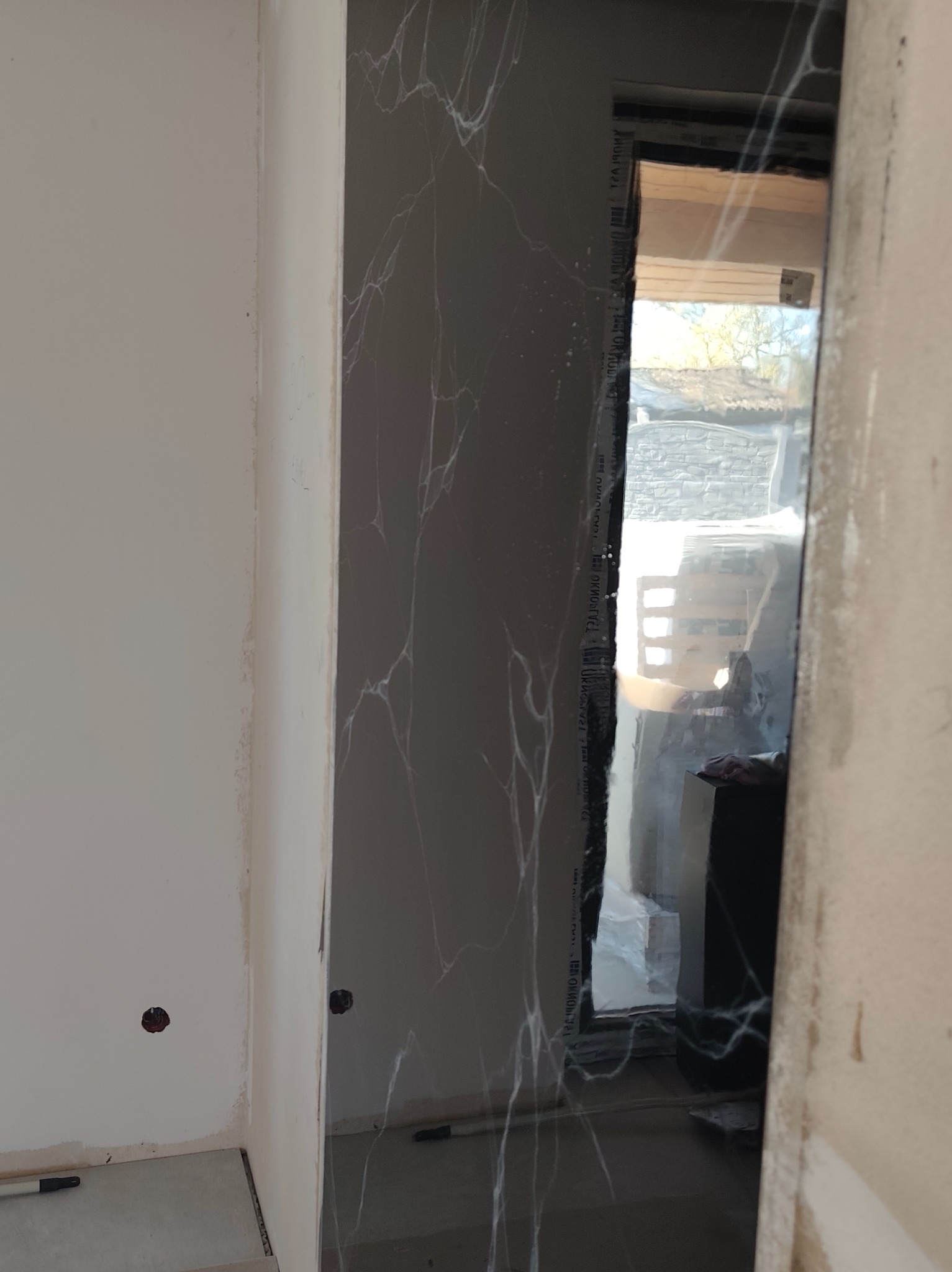 Częściowo wykończona ściana z szarą okładziną imitującą marmur z białymi żyłami, widoczne niedoskonałości na styku z białą ścianą i surowy otwór okienny.