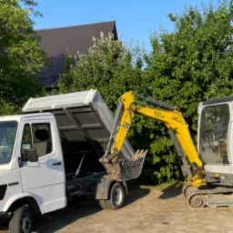 Biała wywrotka Mercedes z podniesioną skrzynią ładunkową obok żółtej minikoparki Wick & Reu.on, na tle zieleni.