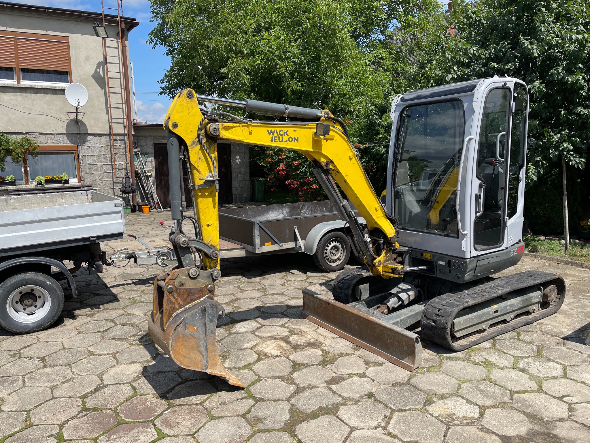 Żółto-szara mini koparka Wacker Neuson EZ28 z łyżką, zaparkowana na brukowanym podwórku, z przyczepką w tle.