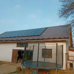Brązowy dach budynku gospodarczego z zamontowanymi panelami fotowoltaicznymi SolarEdge, widoczny fragment trampoliny i podwórka.