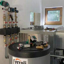 Nowa instalacja grzewcza w kotłowni, widoczne rury, zawory, pompy i zbiornik Ferroli z akcesoriami na pokrywie, małe okno w tle.